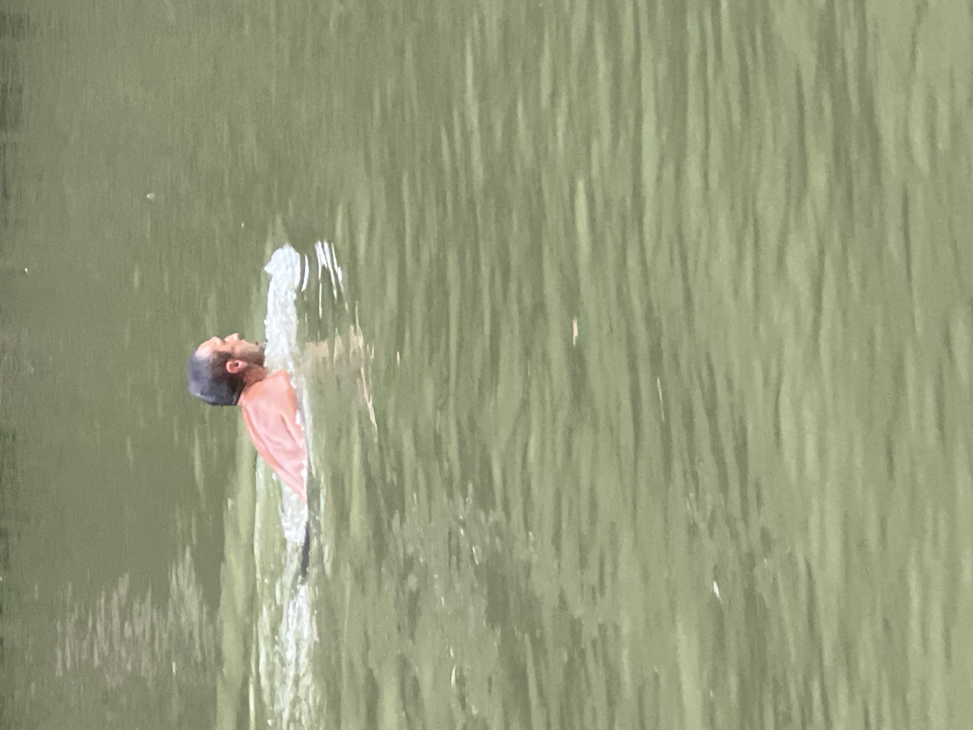 A bald man is swimming in murky green water. He is positioned in the left half of the frame, facing to the right. His back and shoulders are visible above the water, with his head turned slightly to the left. There is a splash of white water around his head and shoulders indicating he is actively swimming. The water has a rippled surface with reflections and light variations. No other people or objects are visible in the water. The environment appears to be a natural body of water.