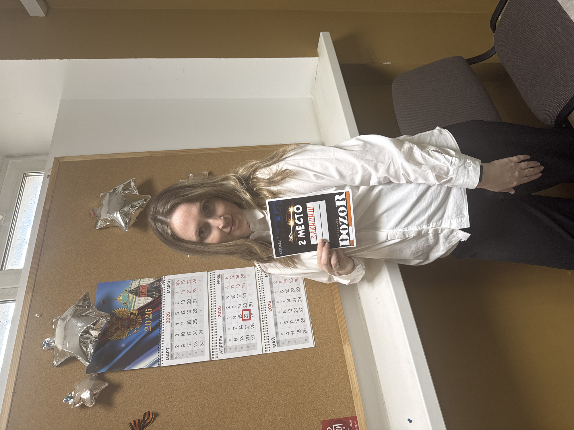 A smiling woman with long blonde hair, dressed in a white button-down shirt and dark pants, is featured in an office-like setting in Chaikovsky, Russia. She holds up a black sign or certificate that prominently displays "АВТОКВЕСТ" (Autogame/Car Quest), "2 МЕСТО" (2nd Place), and "ЗАКИПИШ" (ZAKIPISH), along with "FIGHT GAME" and "DOZOR," indicating an achievement in a game or quest event.
Behind her, a cork board is adorned with celebratory items. A 2026 calendar shows the months of March, April, and May, with the dates April 20, 21, 22, 29, and 30 highlighted in red. The calendar also features an image of a Russian architectural landmark, likely the Kremlin, and the Russian double-headed eagle emblem. Several silver star-shaped balloons and a St. George ribbon, commonly associated with Russian Victory Day, are also pinned to the board.
The background includes a white wall and a brown shelf or partition. A window to the left reveals an overcast day outside. Part of a gray office chair is visible on the right. The overall scene suggests a celebration of a second-place win in an "Autogame Dozor" event, likely around springtime in 2026, given the calendar.