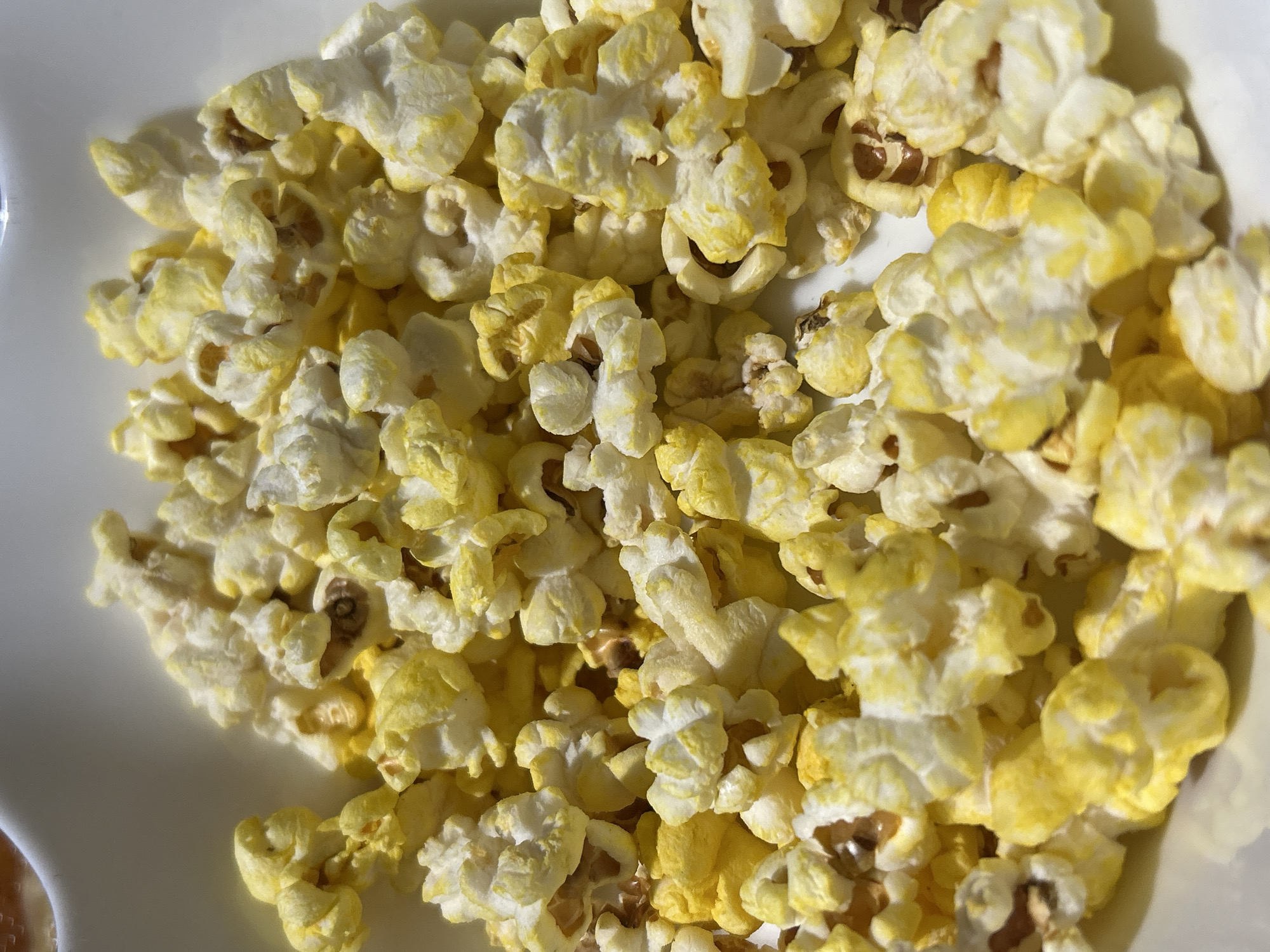 A close-up, overhead shot shows a white bowl filled with popcorn. The popcorn is fluffy and irregularly shaped, with some pieces appearing to be buttered, indicated by a yellowish hue. The lighting is bright, casting subtle shadows. No people or animals are visible. The background is out of focus, with a hint of a curved white edge suggesting the rim of the bowl. The image does not provide any information about the location in the city of Byala Slatina, Bulgaria.