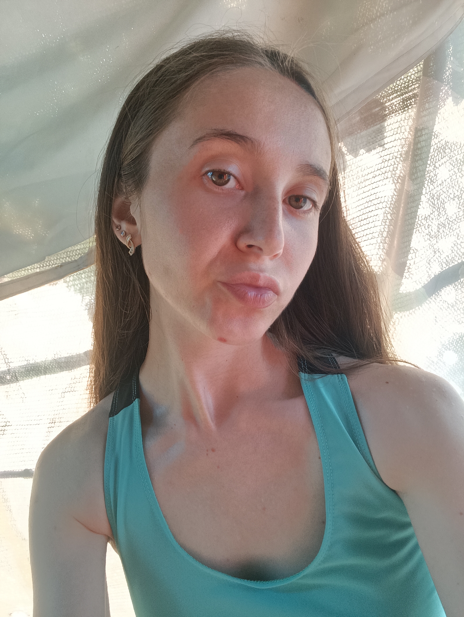 A young woman with long brown hair is taking a selfie. She is wearing a teal tank top. Her face is turned slightly to the left, and she is making a pouty expression. She has a mole on her left cheek and appears to have a small blemish on her chin. She is wearing earrings in her left ear. Behind her, a sheer curtain or screen is visible, with sunlight filtering through it. The image appears to be taken indoors, possibly in a home or a studio. No specific location in Vypasne, Ukraine, can be determined from the image itself.