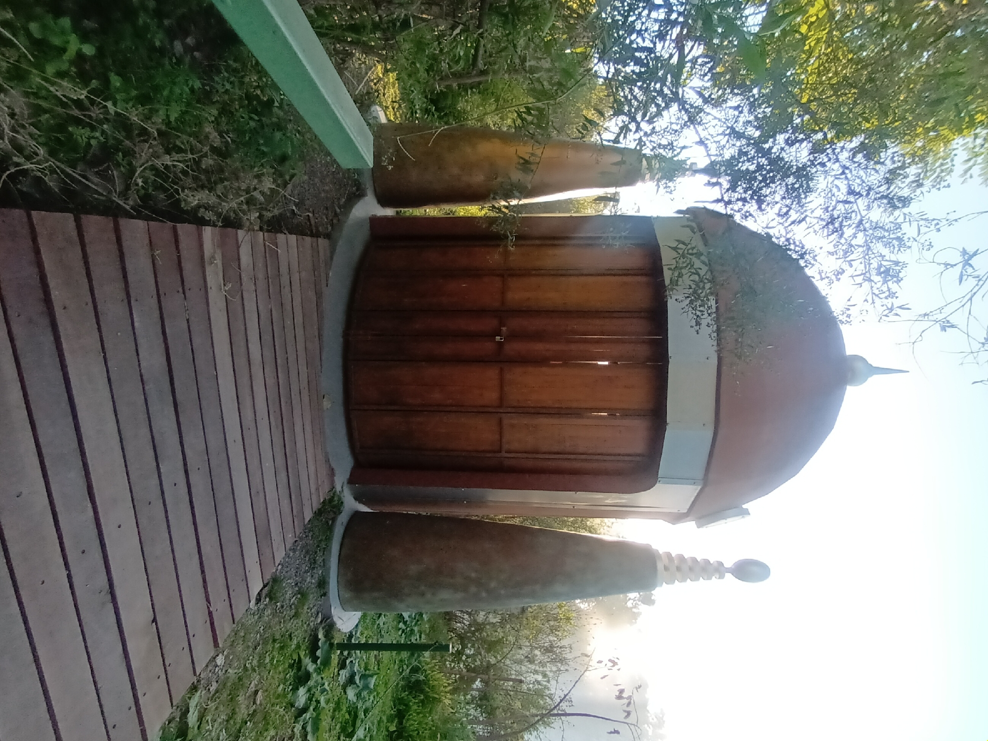 The image shows a wooden structure with a domed roof, resembling a small pavilion or gazebo, situated along a wooden boardwalk. Two large, conical sculptures flank the entrance to the structure. The structure is surrounded by greenery, including trees and shrubs. The location is likely in Buenos Aires, Argentina. There are no people visible in the image. The dominant action is the sun shining through the trees and illuminating the scene. The environment suggests a park or garden setting.