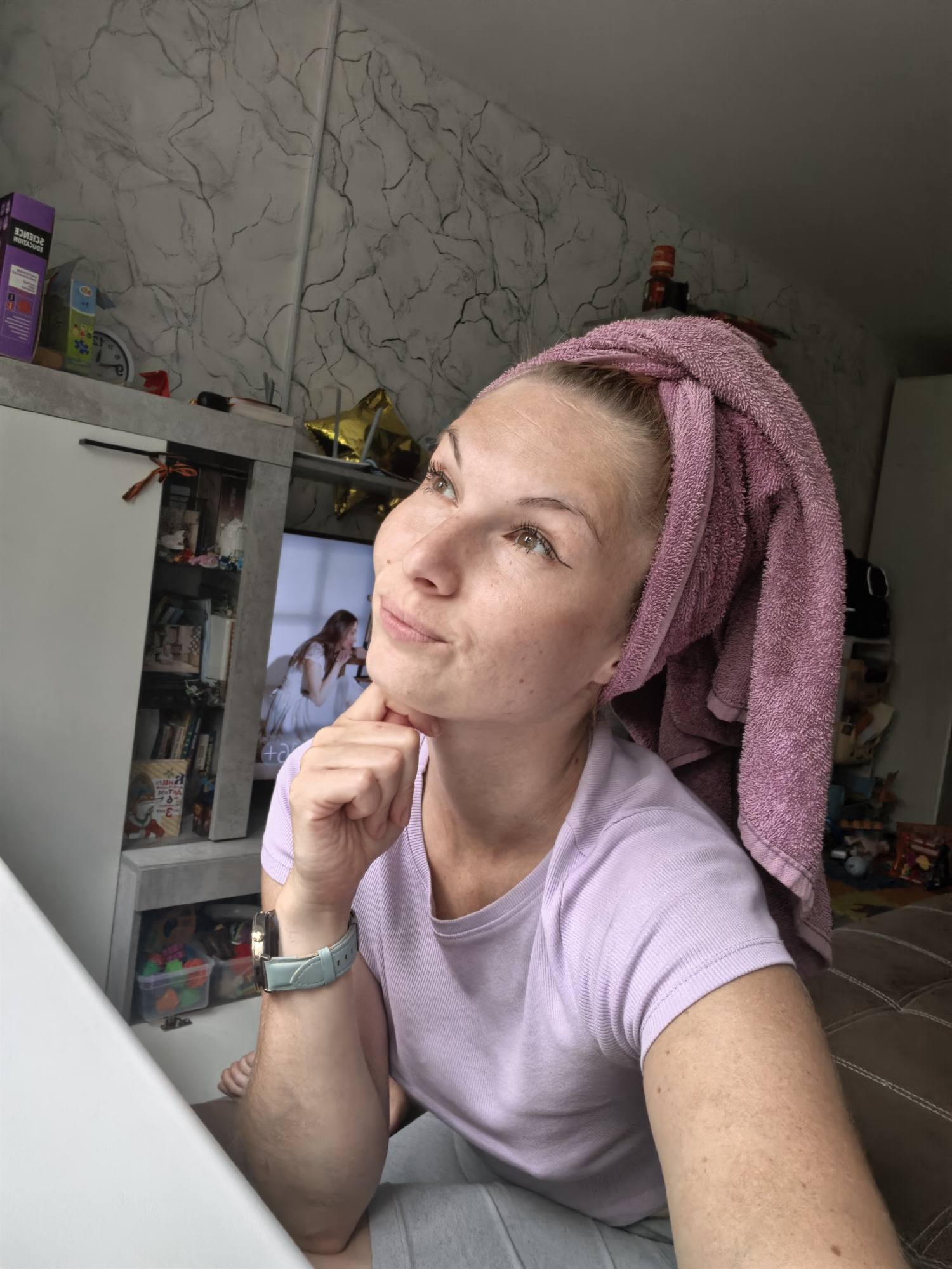 A woman with a pink towel wrapped around her head is looking upwards with her chin resting on her hand. She is wearing a light purple t-shirt and a light blue watch. In the background, there is a shelf unit with various items, a television displaying a scene with two women, and a marbled wallpapered wall. The scene appears to be indoors, possibly in a bedroom or living area. No specific location in Sterlitamak, Russia can be determined from the image.