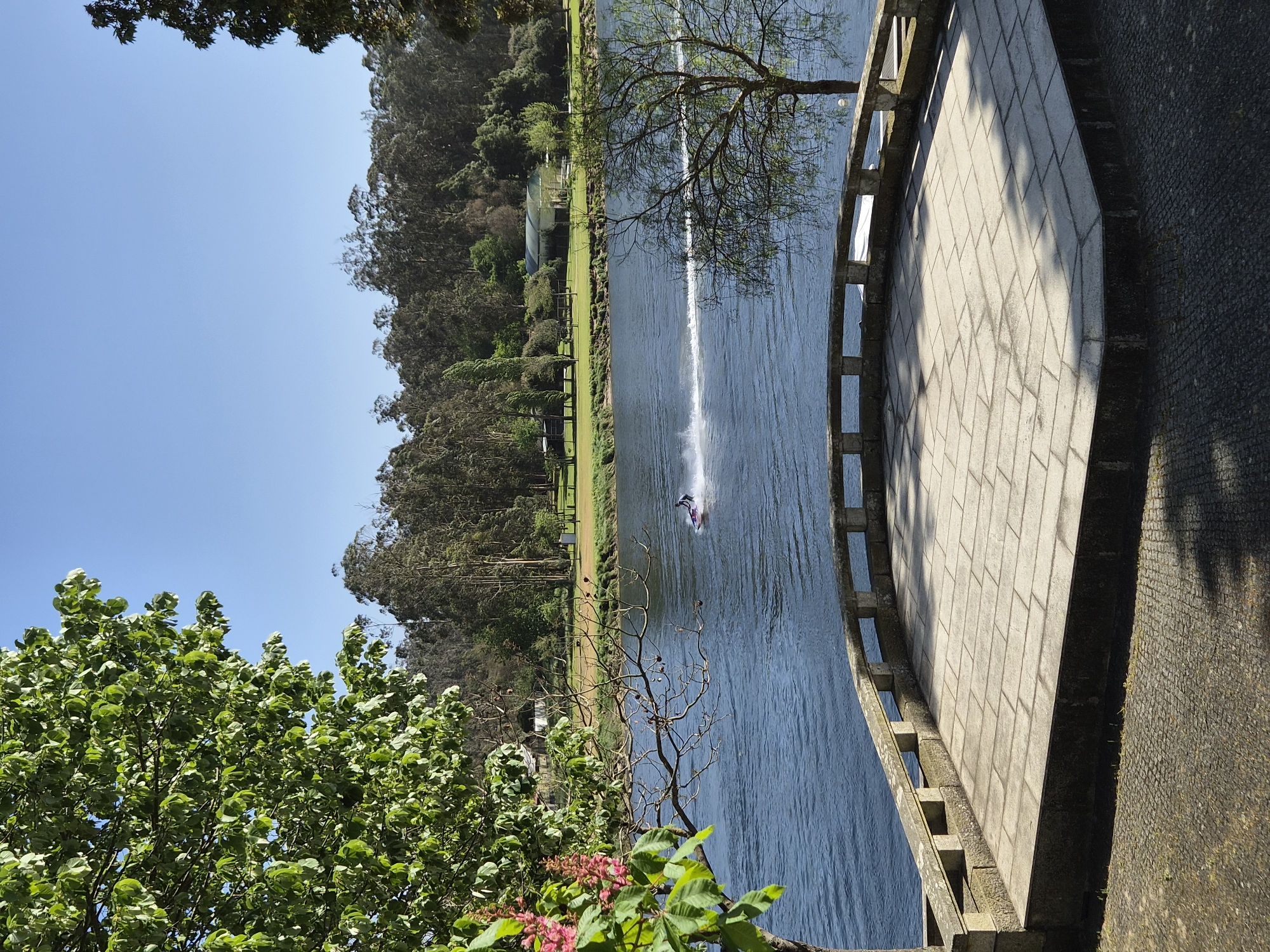 The image shows a view of the Cávado River from a bridge in Esposende, Portugal. A personal watercraft is speeding along the river, leaving a wake behind it. The riverbanks are lined with trees and greenery. A paved walkway or bridge is visible on the right side of the image. The sky is clear and blue.
