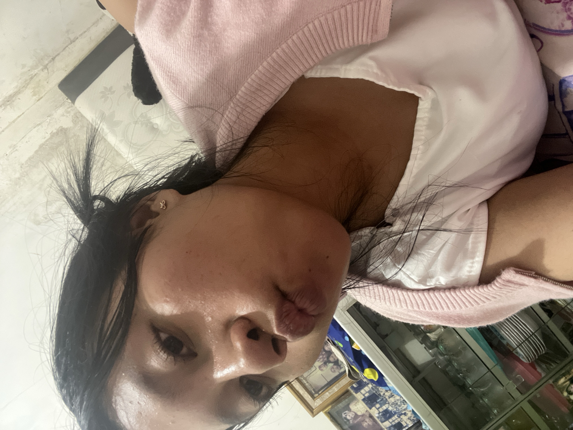 A young woman with dark hair tied back is taking a selfie in what appears to be a bedroom. She is wearing a white t-shirt and a pink cardigan. Her face is close to the camera, and she is pouting her lips. In the background, there is a bed with a patterned mattress and a glass cabinet filled with dishes. A framed picture is visible on the wall to the left. The overall lighting is dim. There is no indication of the location being Pasrukkrajan Satu, Indonesia.