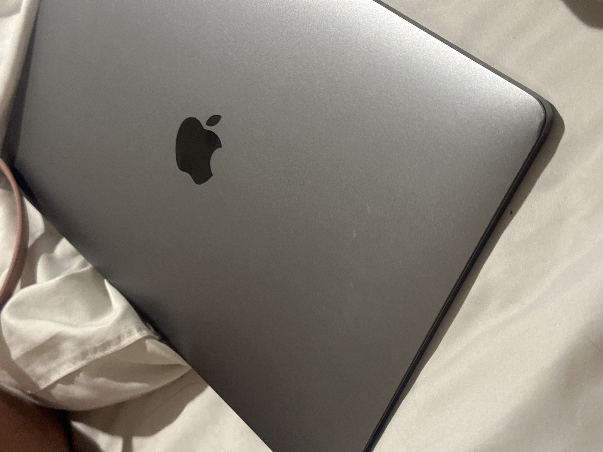 A gray Apple laptop is positioned on a bed covered with wrinkled, light-colored sheets. A small section of what appears to be a light pink strap or garment is visible on the left side of the image. The Apple logo is prominent on the laptop's surface. The location is not discernible from the image alone, and information about North Kuta, Indonesia, cannot be inferred from the provided visual evidence.
