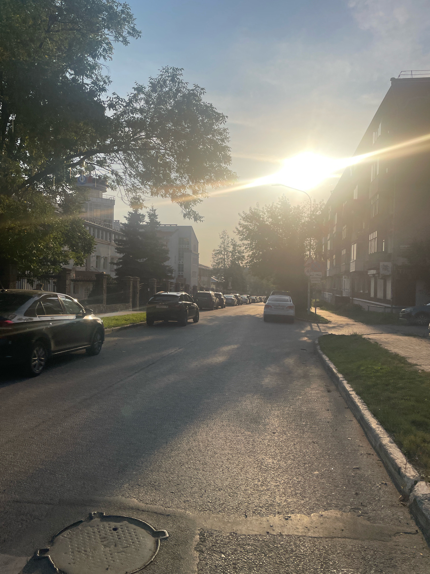 The image depicts a street scene in Izhevsk, Russia, at sunset. The sun is low in the sky, casting long shadows and creating a bright glare. Several cars are parked along the street and some are in motion. Buildings line the right side of the street, and trees are present on the left. The road surface is asphalt, and a manhole cover is visible in the foreground. The overall impression is of a quiet urban environment during the golden hour.