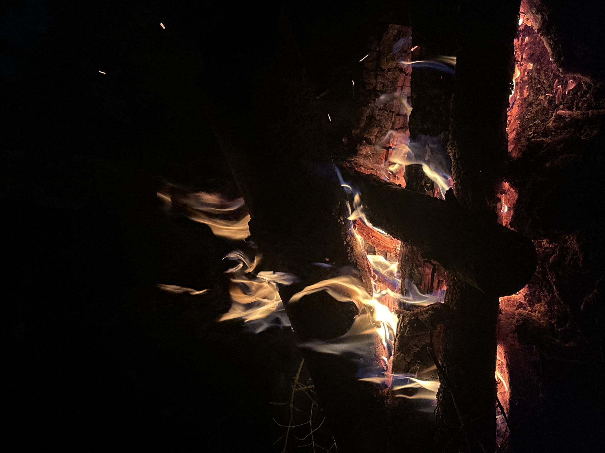 The image shows a nighttime campfire in a dark environment. The fire is burning between several pieces of wood, some of which are silhouetted against the flames. Flames are visible, with a mix of yellow and orange hues and a swirling, ethereal quality. Sparks are visible above the fire. The setting appears to be outdoors.