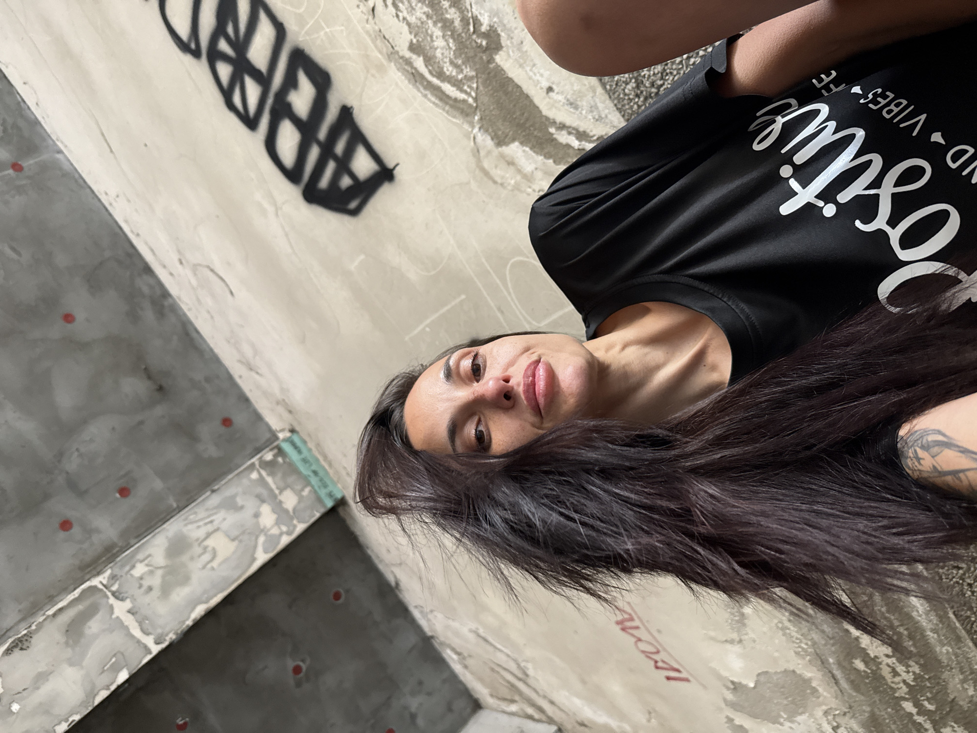 A woman poses indoors against a dilapidated concrete wall marked with graffiti. She wears a black t-shirt with white lettering. The interior space shows signs of disrepair, with exposed concrete and remnants of previous construction or demolition. Red dots are visible on the floor. The location is in an unknown area in Pleven, Bulgaria.