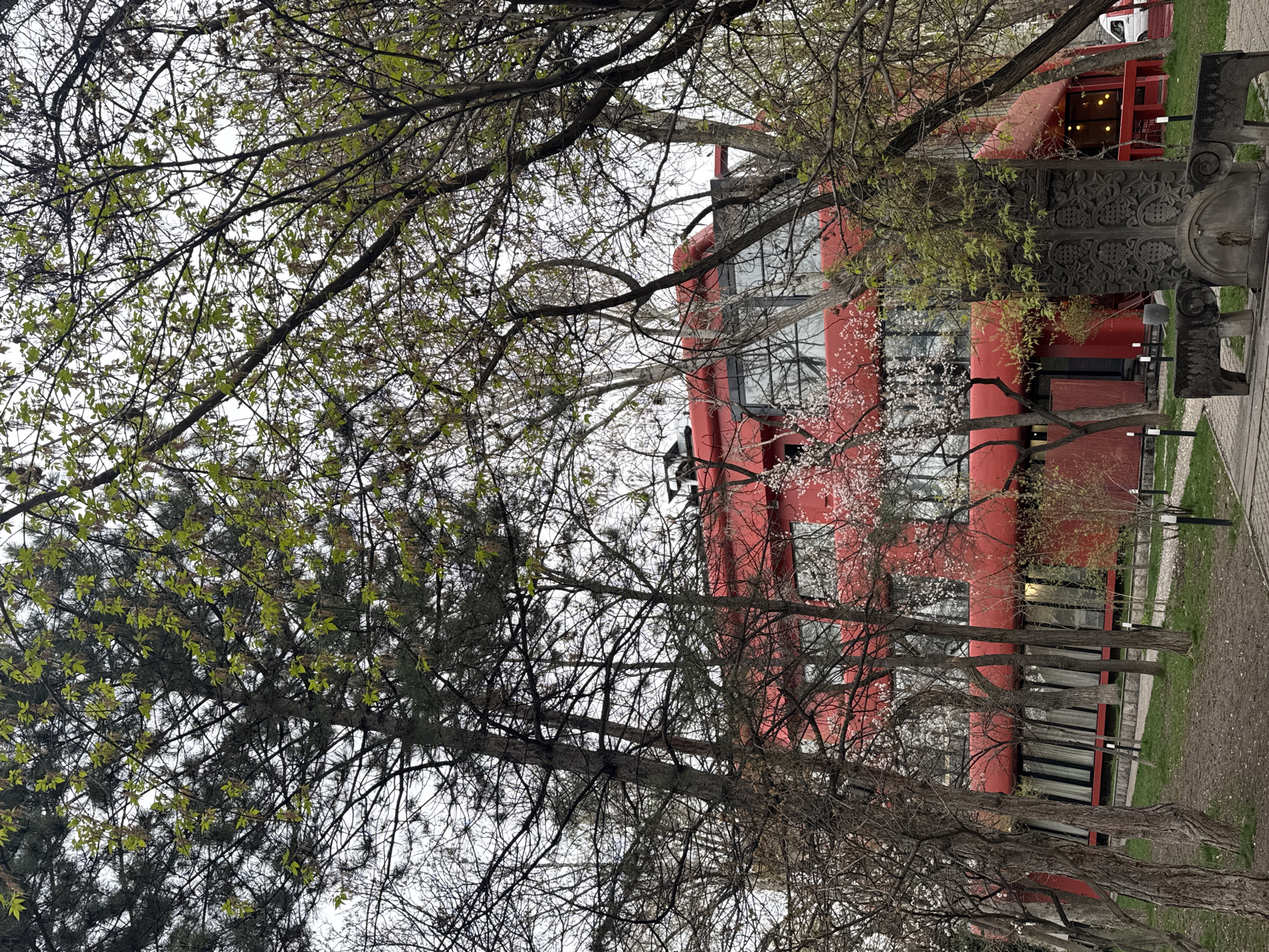 The image depicts a red building framed by trees in Yerevan, Armenia. The building appears to be several stories tall with prominent vertical red sections. Some windows are visible. There's a stone sculpture with intricate carvings partially visible to the right of the building. The trees in the foreground are bare branched, but some have budding green leaves. The ground appears to be a paved walkway or sidewalk and patches of grass. The sky is overcast.