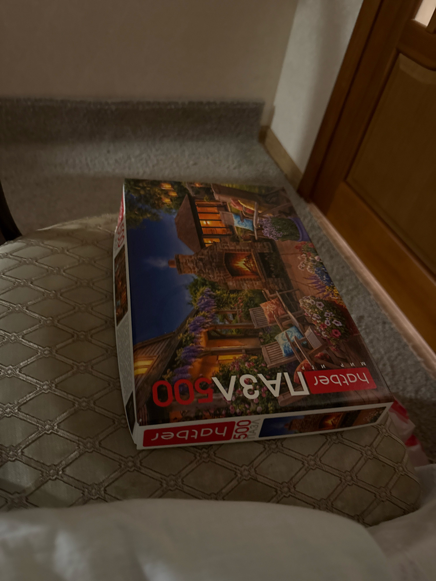 A puzzle box depicting a cozy courtyard scene with a fireplace, chairs, and flowers is resting on a patterned cushion. The puzzle box has red lettering that appears to be in Russian, with "hatber" and "500" visible. The background suggests an indoor setting with a carpeted floor and a wooden doorframe. The image is taken from a high angle, looking down at the puzzle box. There are no people or animals visible in the image. The location is identified as Zavolzhye, Russia.
