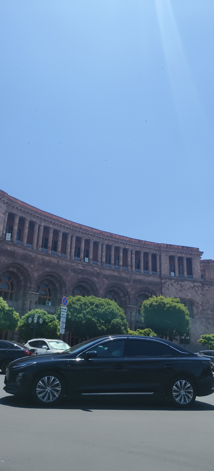 The image captures a daytime scene on a clear, sunny day in Yerevan, Armenia. The main subject is a grand, curved building with a stone facade, featuring multiple levels of arched windows and a colonnade of classical columns on the upper level. The architectural style suggests a historical or significant public building.
In the foreground, a black car is driving on a paved road. The car is sleek and modern, with tinted windows and distinctive alloy wheels. To the left of the car, another white SUV is partially visible. Behind the cars and in front of the building, there are lush green trees, providing a contrast to the stone architecture. A street sign with multiple panels and symbols, indicating traffic regulations, is also visible.
The sky is a bright, clear blue, with a few small birds or insects scattered across it, suggesting it might be a warm day. The sunlight casts shadows, indicating the time of day is likely midday or early afternoon. The overall impression is one of urban life in a historic city, with modern vehicles navigating the streets in front of impressive architecture, under a pleasant, clear sky.