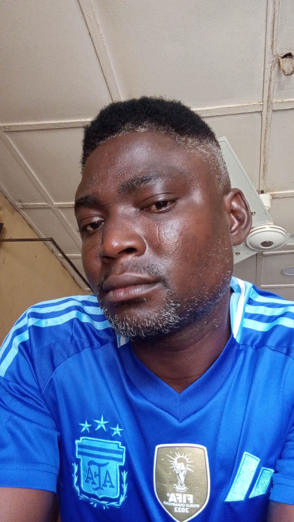 A dark-skinned man is captured in a close-up, chest-up portrait shot. He has short, dark, curly hair with visible grey or white strands at the temples and sideburns, extending into a short beard with similar grey highlights. His face appears moist or shiny, possibly from sweat, and he has a neutral to slightly pensive expression, looking downwards and to the viewer's left. A faint, vertical scar or mark is visible on his left cheek (viewer's right).
He is wearing a vibrant blue sports jersey featuring light blue stripes on the shoulders and sleeves. On the left chest of the jersey, a crest is embroidered with the letters "AJA" inside a shield-like emblem, surrounded by laurels, and topped with three white stars. On the right chest, a gold patch with the FIFA World Champions 2022 logo is visible. The text on this patch, "FIFA® WORLD CHAMPIONS 2022," appears mirrored, suggesting the image is likely a selfie or has been flipped horizontally.
The setting is indoors, with a light-colored, possibly paneled ceiling visible in the background. A white ceiling fan with brown accents is partially visible in the upper right corner. The overall impression is a casual, indoor environment. The image was taken in Sabon Tasha, Nigeria.