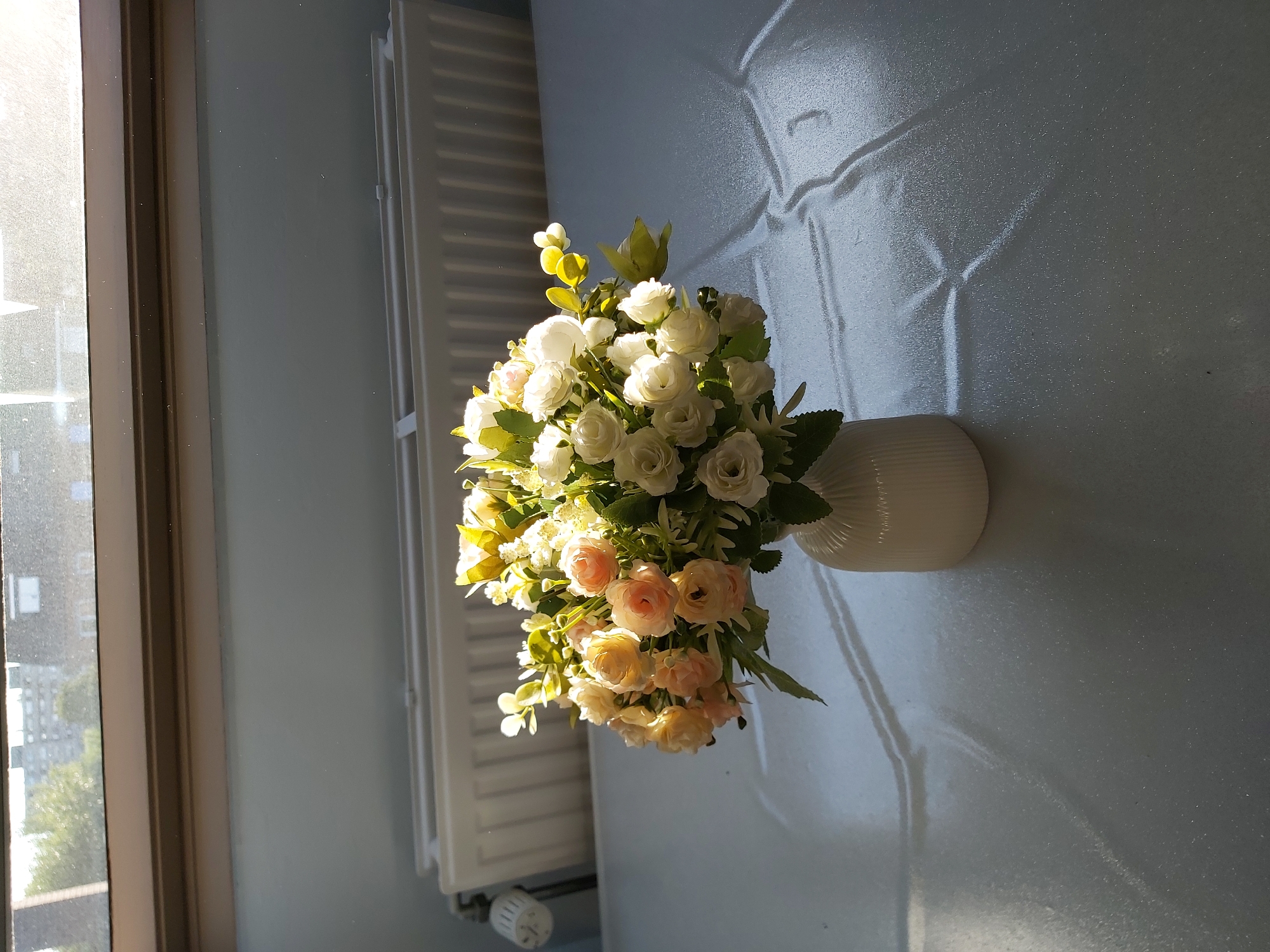 A vase of artificial flowers sits on a textured surface, casting shadows. The flowers are a mix of white and pale pink roses, with green foliage. The vase is white and ribbed. Behind the vase, a radiator is visible against a light blue wall. Sunlight streams in from a window, indicated by the bright illumination and the patterns of light and shadow on the surface. The scene appears to be indoors, possibly in a room with a view of buildings and trees through the window.