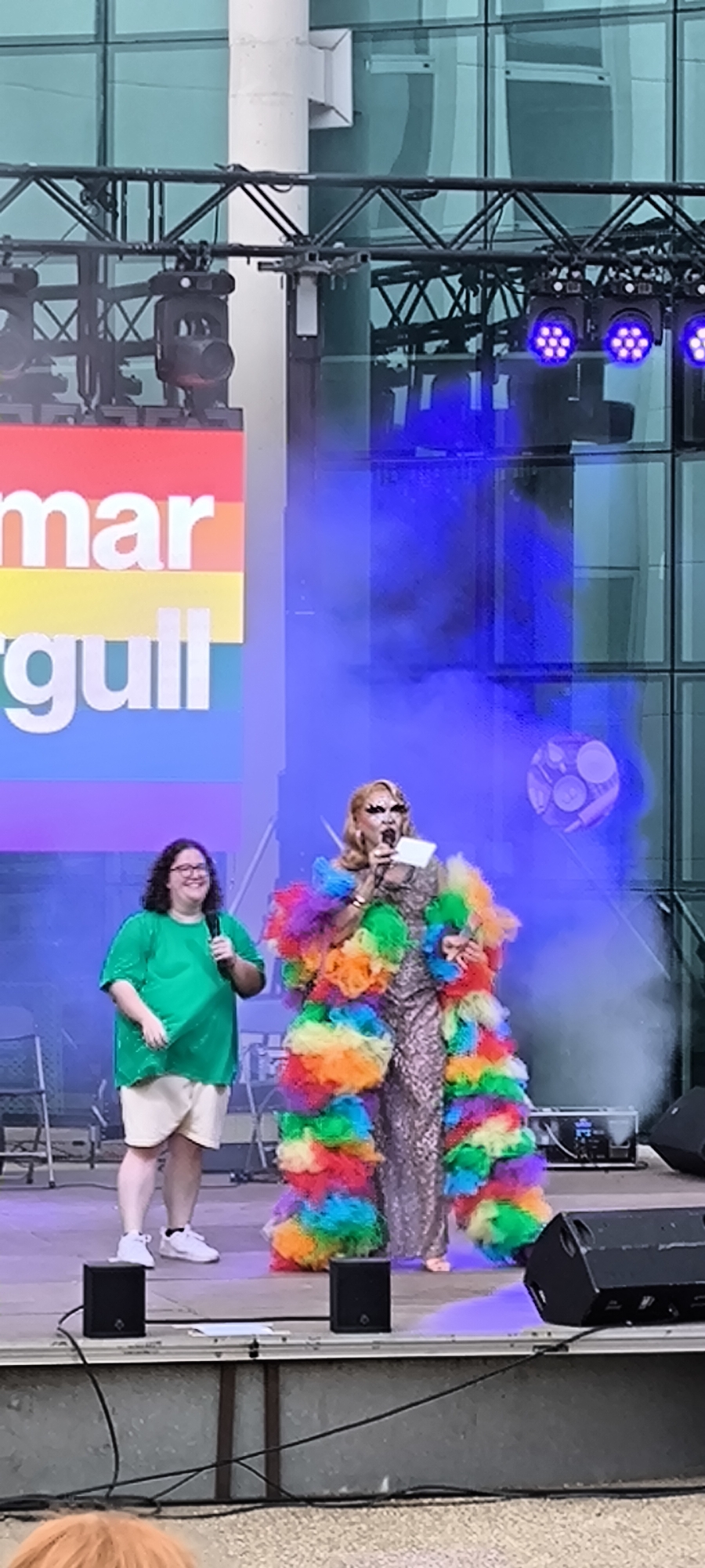 Two individuals are on a raised stage in front of a building with a glass facade. The individual on the left is wearing a green t-shirt, khaki shorts, and white sneakers, and is holding a microphone. The individual on the right is dressed in a sparkly silver dress and a vibrant, multi-colored furry coat. They are also holding a microphone and appear to be speaking. Behind them, a large banner with rainbow stripes and white text reads "mar orgull". Stage lighting, including blue spotlights, is visible. The event appears to be taking place outdoors, possibly in the Plaça de la Vila in Ripollet, Spain, given the context provided by the banner, which suggests it is related to Pride celebrations. The atmosphere is festive, with smoke effects adding to the visual presentation.