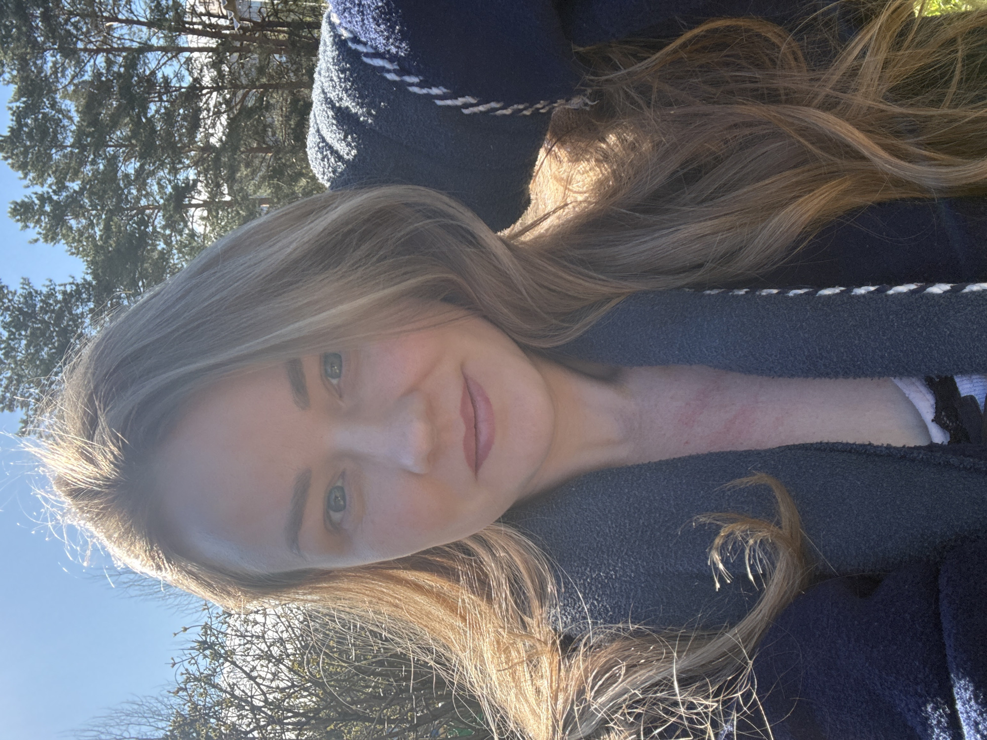 A young woman with light brown hair, a fair complexion, and blue eyes poses outdoors, likely in Chaikovsky, Russia, given the prompt. She wears a dark blue coat with a light gray and white trim. The background includes bare trees against a clear blue sky, suggesting a late autumn or winter season. The lighting is bright, and the focus is on the woman's face.