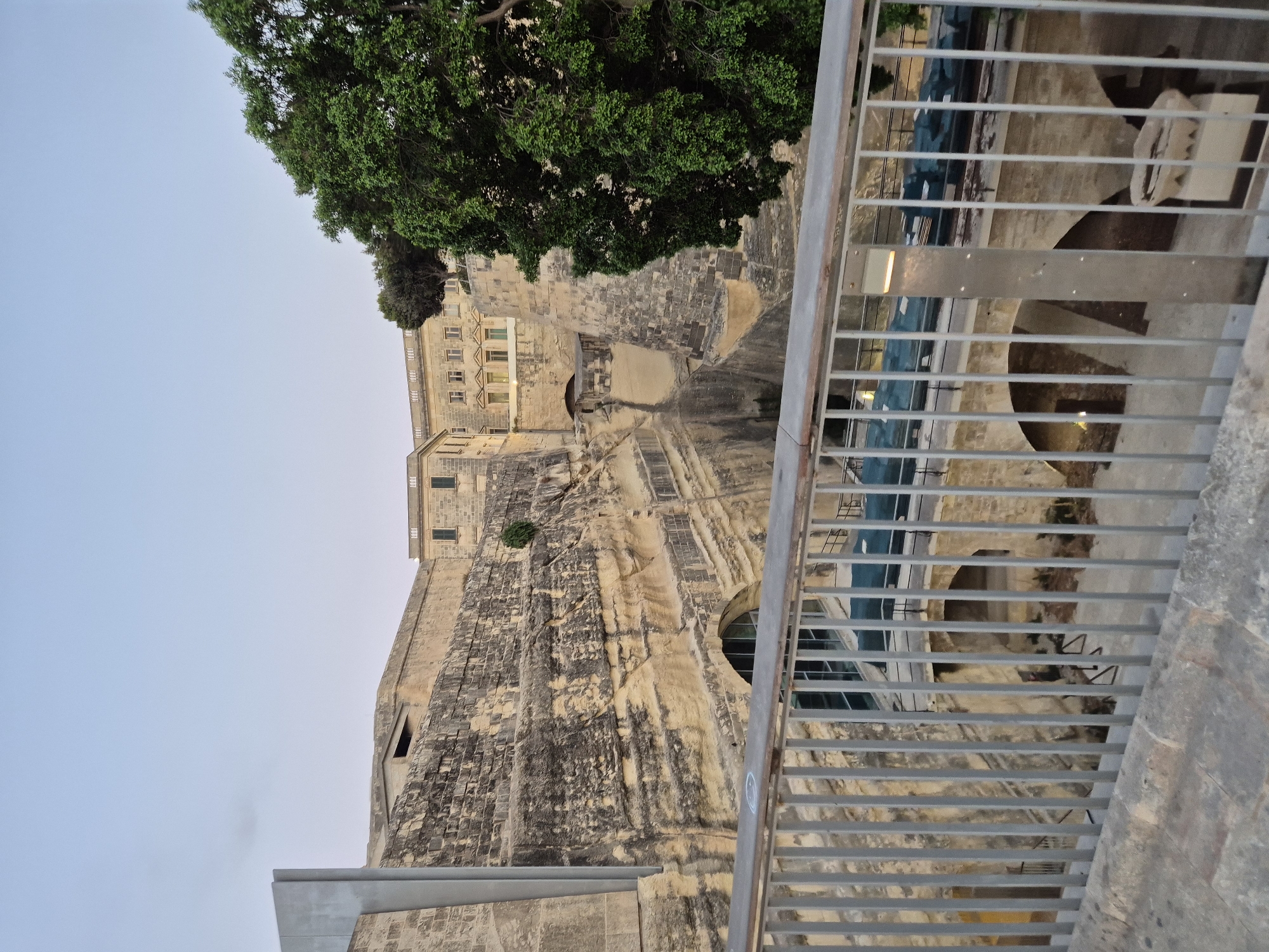 The image depicts a view of the Valletta City Gate complex, specifically the area surrounding the ditch and fortifications. A metal railing in the foreground separates the viewer from a large, stepped rock-cut ditch. Beyond the ditch, ancient stone fortifications rise steeply, featuring layered stonework and archways. A modern, grey metal structure is visible on the left side of the fortifications. At the top of the rock face, a light-colored building with multiple windows and a patterned facade is visible, partially obscured by trees. A large, lush green tree dominates the right side of the frame, its branches extending over the fortifications. The sky above is a pale, even blue, suggesting early morning or late evening. Several arched openings and structures are visible within the ditch, hinting at historical passages or chambers. A modern walkway or bridge, possibly with lighting, spans across the ditch. The overall impression is one of historic architecture integrated with natural rock formations, set against a serene sky.