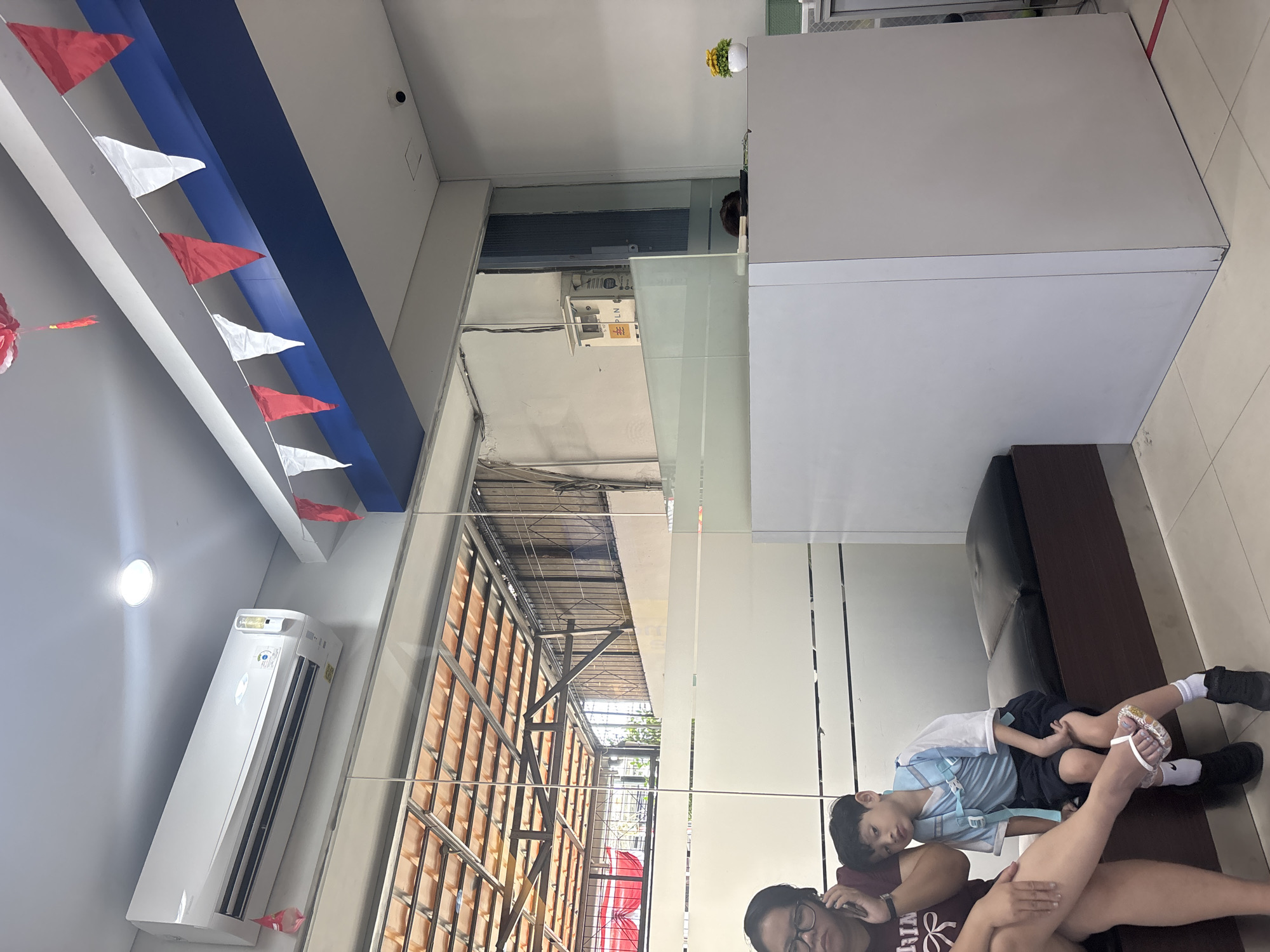 In North Jakarta, Indonesia, a mother and son are seated on a dark brown bench in what appears to be a waiting area or reception. The mother, wearing a maroon t-shirt and glasses, is on her phone. The son, in a blue and white school uniform with a backpack, is leaning his head on his mother's shoulder, looking sleepy. A white air conditioning unit is mounted on the wall above them. Red and white triangular flags are strung across the ceiling, suggesting a festive occasion. A frosted glass partition separates the waiting area from a counter area, where a small yellow flower in a white pot sits on a white counter. Reflections in the glass show a tiled roof structure outdoors. A security camera is visible on the ceiling in the background.