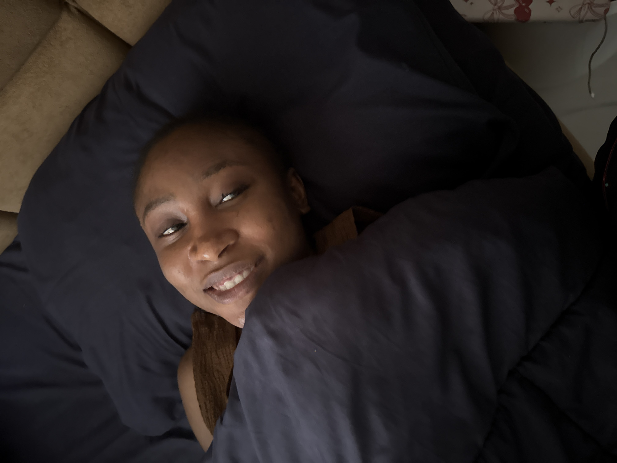 A medium shot, taken from a high angle, shows a young Black woman lying in bed, covered by a dark blue comforter. She is facing the camera and smiling, with her head resting on a dark blue pillow. The woman has her eyes open and is wearing a brown top. The background shows a brown couch and a section of a wall with a decorative pattern. The scene appears to be indoors, likely in a bedroom. No specific landmarks of Abuja, Nigeria are visible.