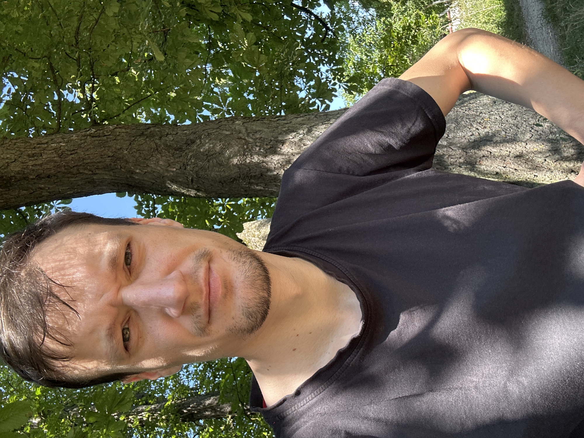 This is a selfie taken outdoors in Makiv, Ukraine. The subject is a white male with brown hair and a short beard, wearing a black t-shirt. He is standing in front of a large tree with green foliage. The background shows more trees and a clear blue sky. The lighting suggests it is daytime.
