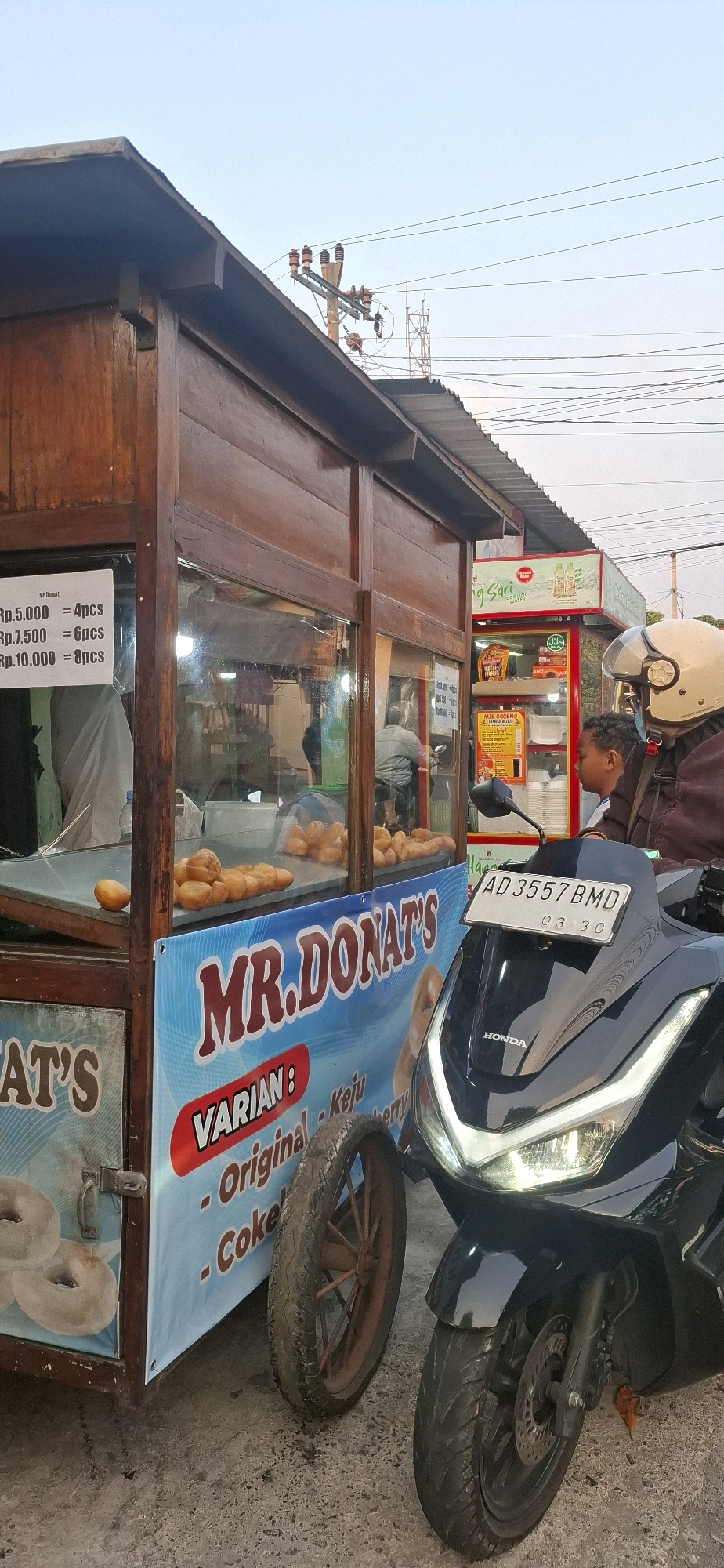 In Desa Gagaksipat, Indonesia, a roadside donut stand named "Mr. Donat's" is captured in a medium shot. The stand, constructed of wood and featuring glass display cases, is adorned with a blue banner showcasing the business name and a list of donut varieties such as "Original," "Keju" (Cheese), and "Cokelat Berry" (Chocolate Berry). Displayed on the stand are rows of golden-brown donuts. A white sign with pricing, indicating "Rp.5.000 = 4pcs", "Rp.7.500 = 6pcs", and "Rp.10.000 = 8pcs", is visible. A black scooter, with its headlight illuminated and license plate "AD 3557 BMD", is parked in front of the stand. A person wearing a helmet is seated on the scooter, partially visible. To the right of the donut stand, another small vending booth is visible, displaying a sign for "Mie Goreng" (Fried Noodles). The background shows power lines and utility poles against a clear sky. The ground is paved with asphalt.