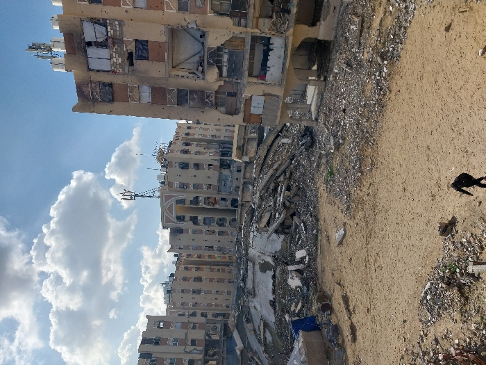 The image displays a scene of widespread urban destruction in Khan Yunis, Palestinian Territory, under a sky with scattered white clouds. Multiple multi-story concrete buildings, appearing to be residential blocks, exhibit severe damage, including large sections of collapsed walls, exposed interiors, numerous projectile impact holes on their facades, and shattered or missing windows, some of which are covered with makeshift materials. The structure in the immediate right foreground shows significant damage to its upper and lower floors. In the mid-ground, a substantial pile of rubble and collapsed building materials indicates a completely destroyed structure. The ground throughout the foreground and mid-ground is largely bare, disturbed sandy soil interspersed with extensive debris from destroyed buildings, including concrete fragments and twisted rebar. A single individual, dressed in dark clothing and carrying a backpack, walks across the sandy ground in the lower foreground, moving away from the viewer towards the central rubble area. Communication towers with multiple antenna arrays and water tanks are visible on the rooftops of the less-damaged buildings in the background.