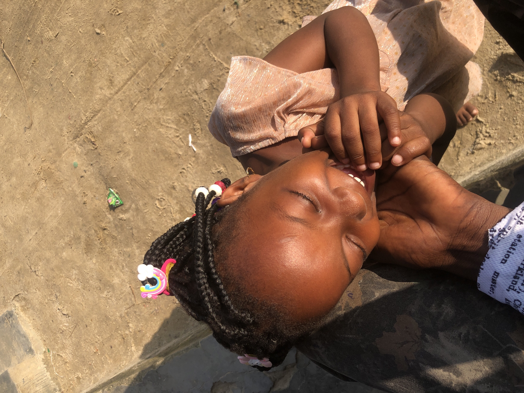 An overhead, high-angle, medium shot captures a young Black girl, with her eyes closed and mouth open in laughter, her hands clasped. She has her hair styled in braids and decorated with colorful hair clips shaped like rainbows and flowers. She is wearing a shimmering peach-colored dress. The girl is being held by an unseen individual, whose hand is visible holding her own hands. The background is composed of rough, sandy ground with some scattered debris. The scene suggests a moment of joy and connection, possibly outdoors on a sunny day. Given the context provided, the location is implied to be Ado, Nigeria.