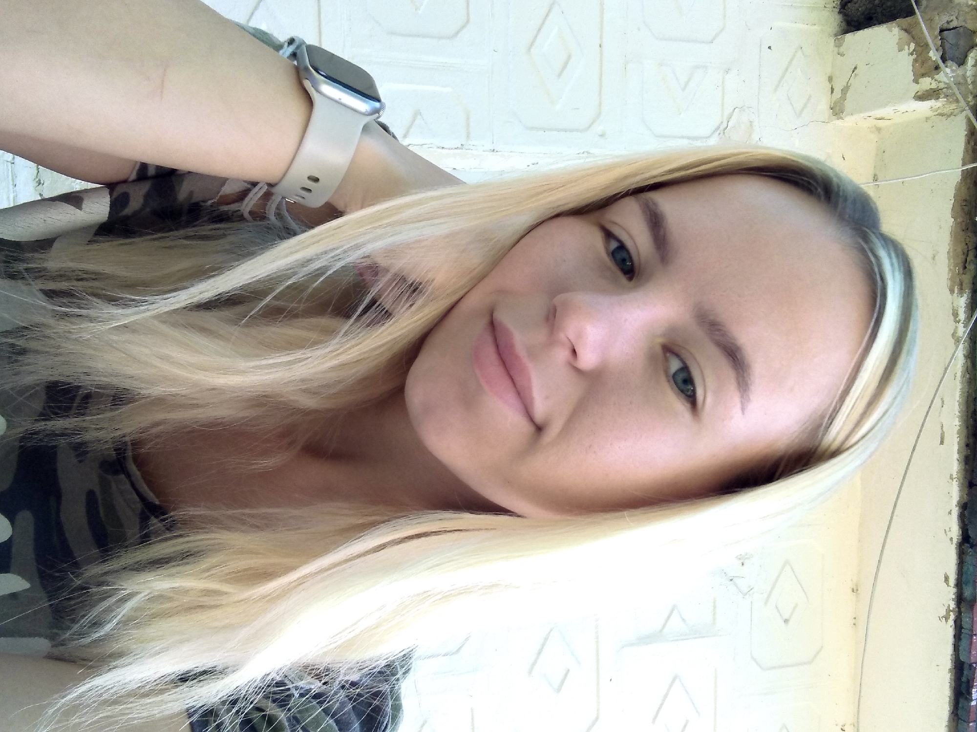 In Radisne, Ukraine, a young woman with blonde hair is captured in a close-up, head-and-shoulders portrait against a white, textured wall with a rough concrete overhang at the top. She is wearing a camouflage t-shirt and a light-colored smartwatch on her left wrist. Her head is tilted, and her left hand is resting on her cheek. The lighting suggests an outdoor, daytime setting.