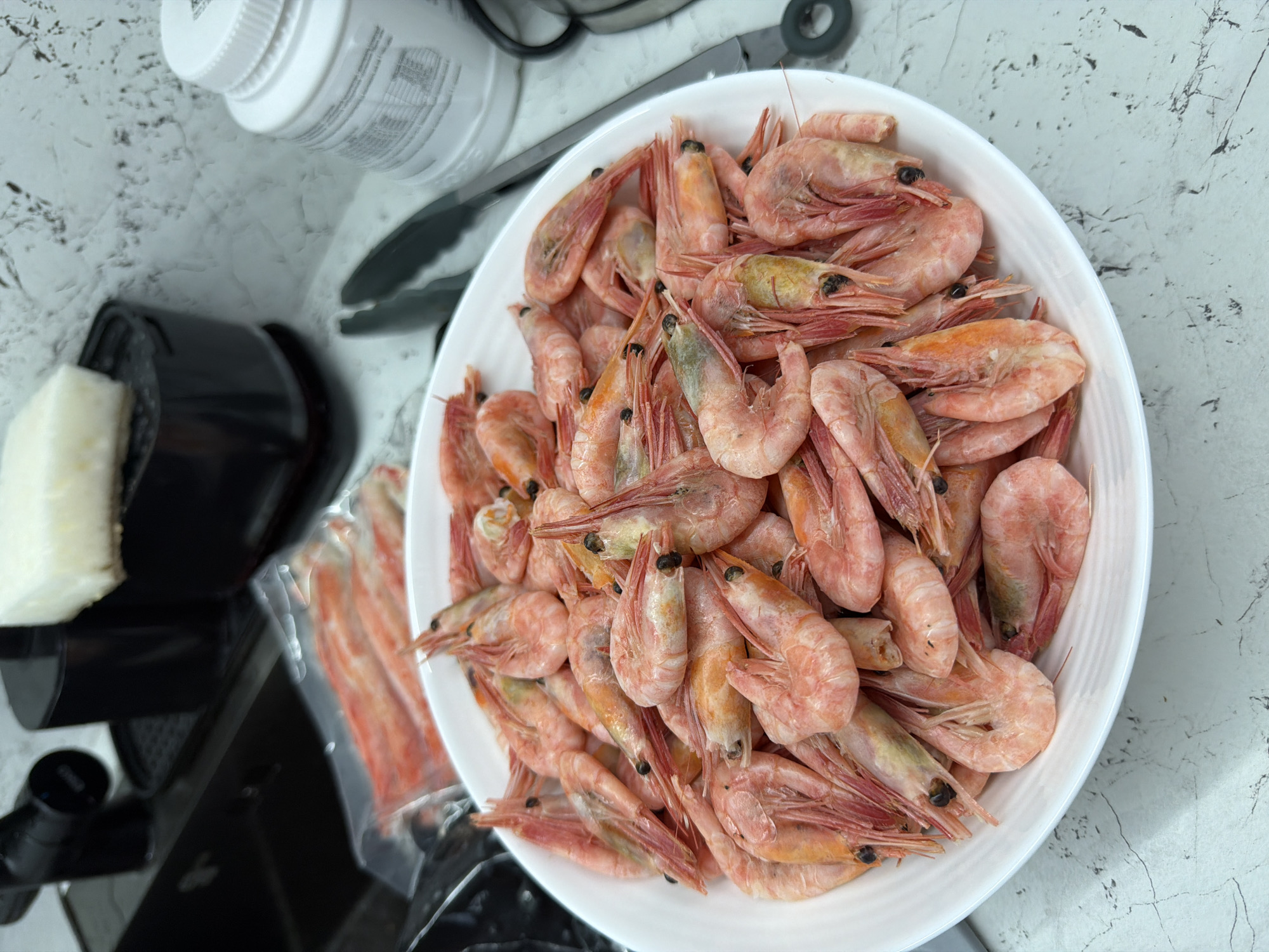A close-up, overhead shot shows a white bowl filled with cooked shrimp, piled high. The shrimp are pink and orange with visible legs and antennae. The bowl rests on a light-colored, speckled countertop. In the background, out of focus, are various kitchen items including a black appliance, a white rectangular object, a clear plastic bag containing more pink seafood, and a white plastic container. The scene does not provide any identifiable landmarks or features specific to Novorossiysk, Russia.