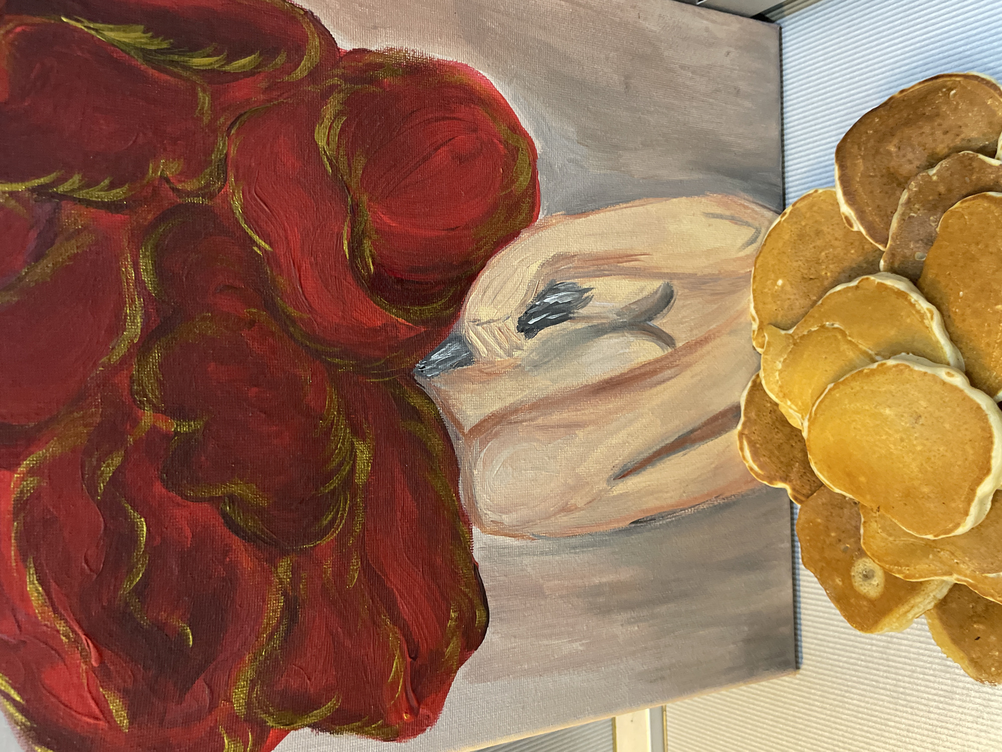 The image displays a still life composition featuring a painting positioned vertically in the background and a pile of pancakes in the foreground. The painting depicts an abstracted human figure, likely female, from the chest up. The figure's head is tilted backward and largely obscured by a mass of voluminous, textured red forms, outlined with streaks of golden-yellow, which represent hair or a symbolic element. The visible skin tones are rendered in warm beige and peach hues. A dark, irregular mark is present on the upper chest/neck area of the painted figure. The background within the painting is a flat, light grey. The canvas itself exhibits a visible texture. In the foreground, a stack of approximately fifteen to twenty small, circular pancakes is arranged on a flat, white surface with fine horizontal ridging. The pancakes vary in color from light golden-brown to a darker brown, suggesting slight differences in cooking. The specific location in the city of Sochi, Russia, cannot be determined from the visual information provided in the image.