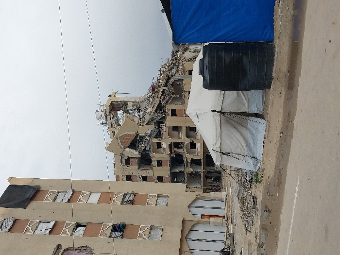 A damaged building stands prominently in the frame, showing structural devastation with missing walls and rubble. Adjacent to it is a relatively intact building with some damaged windows. A white tent and a large black water tank are situated in front of the buildings, possibly indicating temporary shelter and resource provision. The scene suggests an aftermath of conflict or disaster, with a gray sky overhead contributing to the somber mood.