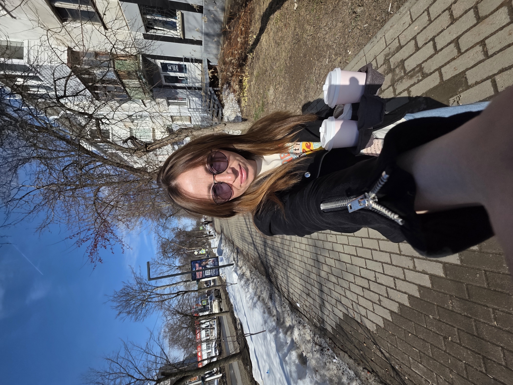 A forensic examination of the image reveals a female subject standing on a paved sidewalk in Chaikovsky, Russia, during what appears to be early spring or late winter.
**Subject:** A single adult female is positioned centrally, holding the camera in a selfie orientation, extending her right arm forward. She has long, light brown hair and wears dark, round-framed sunglasses. Her attire includes a black zip-up jacket with a prominent silver zipper pull on the right sleeve, and a light-colored t-shirt with visible graphics. Her expression is neutral as she looks directly at the camera. In her left hand, she holds a dark-colored tray containing two light pink cylindrical cups with white lids, consistent with takeaway beverages.
**Foreground/Midground (Left):** The paved sidewalk, constructed from rectangular gray pavers, extends into the distance. To the left of the sidewalk is a strip of melting snow and exposed earth. Beyond this, a road is visible with several vehicles in the distance. The area is lined with numerous bare deciduous trees, some exhibiting reddish buds on their branches. Streetlights and billboards, including one displaying a person, are present along the roadside.
**Foreground/Midground (Right):** To the subject's immediate right, a patch of dry earth contains withered vegetation and sparse emerging yellow flora. Behind this, a multi-story building with a light-colored exterior (likely white or light gray) features multiple windows and balconies, one of which has a faded green lower panel. An external staircase leads to an entrance door displaying signs. More bare trees are interspersed within this area.
**Background:** The sky is clear blue with minimal cloud cover, and a faint white contrail from an aircraft is visible. The overall scene is illuminated by direct sunlight, casting distinct shadows.