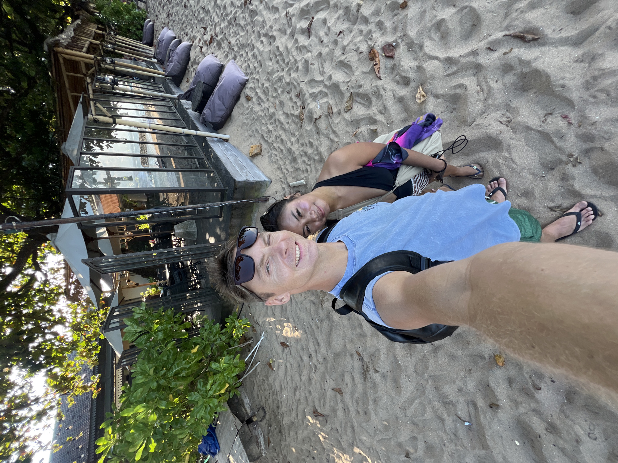 A high-angle, full-shot selfie captures a young Caucasian man and a young Asian woman on a sandy beach in Denpasar, Indonesia. The man, with short brown hair and sunglasses, faces the camera, smiling broadly and holding the phone. He wears a light blue t-shirt, green shorts, and a black backpack. The woman, with dark hair, is positioned slightly behind and to his right, leaning her head on his shoulder and smiling softly. She wears a black halter-neck swimsuit and is holding a purple and white sarong. They are standing on a sandy beach with scattered leaves and debris. In the background, a row of grey beanbag chairs sits on the sand in front of a modern structure with glass walls and a thatched roof, likely a beachside restaurant or cafe. Lush green trees with dappled sunlight filter through their leaves, casting shadows on the sand and the building. The scene suggests a relaxed atmosphere, possibly a vacation or a casual outing at a beach resort.