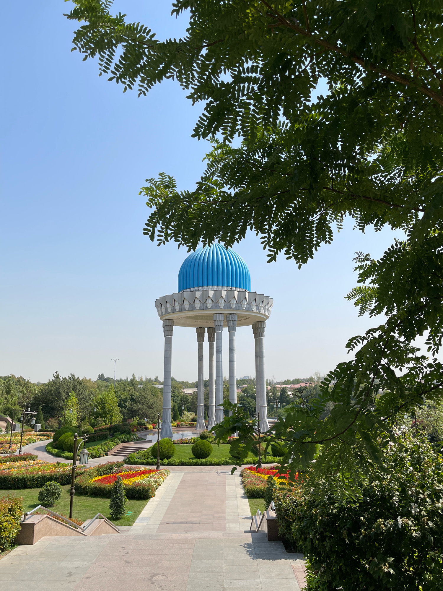 This is a photograph of the Independence Monument in Tashkent, Uzbekistan. The monument features a large blue dome supported by several tall, white columns, set against a clear blue sky. The foreground is a pathway leading towards the monument, with meticulously manicured gardens on either side, filled with colorful flowers and lush green bushes. Trees with dense foliage partially obscure the view, framing the scene. The pathway is paved with large, light-colored tiles. The overall impression is one of a well-maintained public space bathed in daylight.