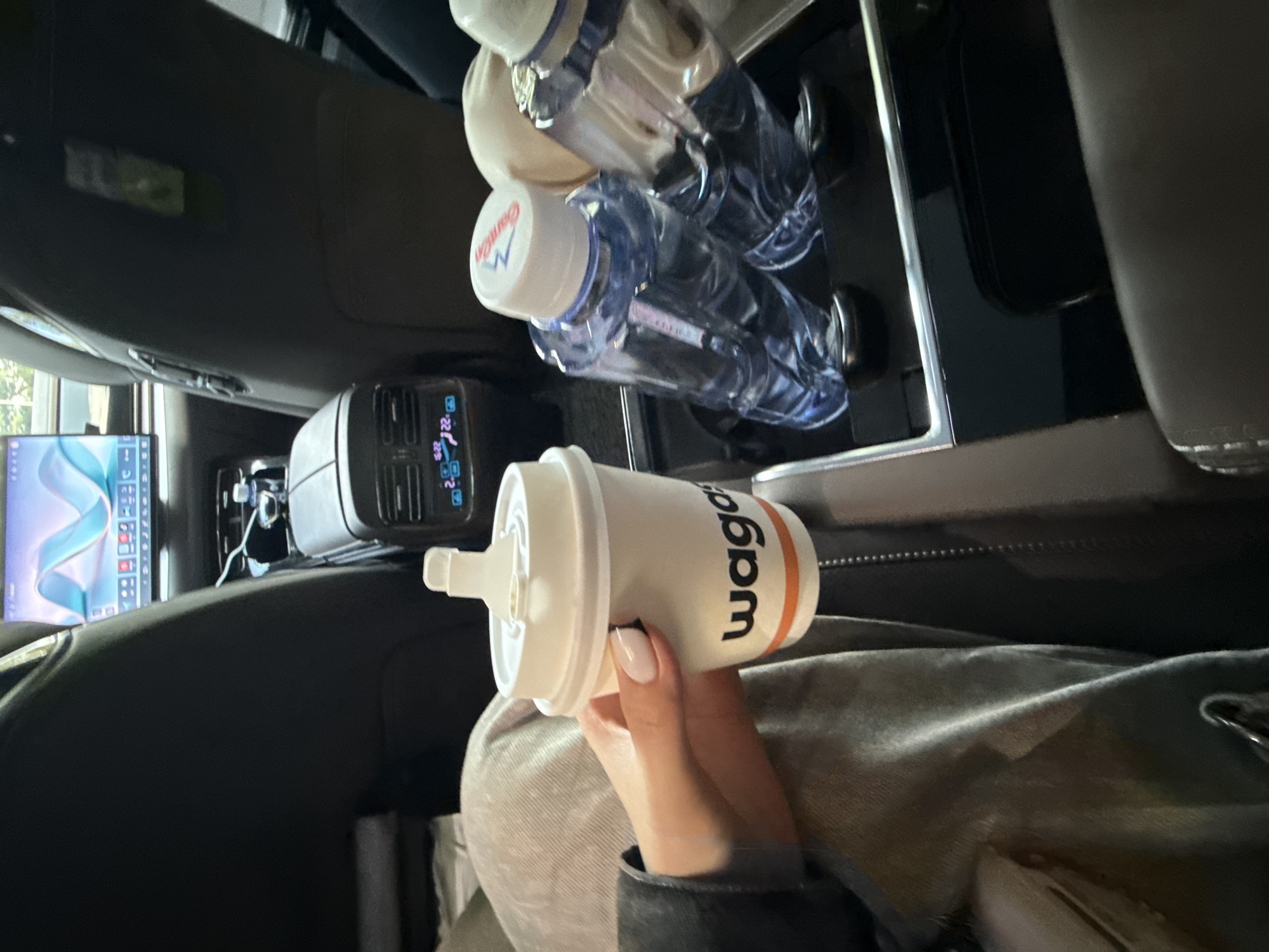 The image captures a scene inside the passenger cabin of a vehicle, likely a car, during daylight hours. The perspective is slightly angled and vertical.
The central focus is a person's right hand, adorned with light-colored nail polish, holding a white, cylindrical reusable cup. The cup features a white lid with a raised straw mechanism. On the side of the cup, the brand name "wago" is clearly visible in black letters, with the 'g' and 'o' set against an orange background stripe.
In the background, on the right side of the frame, two clear plastic water bottles are positioned in what appears to be a cup holder or storage area. Both bottles have blue caps; the cap of the lower bottle displays the brand name "Cambon" in white text. The surrounding car interior is dark, featuring black and dark grey upholstery and shiny, possibly metallic or plastic, trim.
To the left of the cup, a black portable power station or inverter is visible. Its digital display shows readings such as "22" and "20," along with icons suggesting battery status and power output. Further to the left, a digital screen, potentially a car infotainment display or a mounted tablet, shows a dynamic blue and white abstract graphic, possibly a map or a digital wallpaper, with several small application icons visible along its bottom edge.
The scene suggests a journey or travel within the vehicle, emphasizing convenience and personal items. The overall mood is calm and practical. No specific emotions are discernible. The location context for this scene is Haizhu District, China.