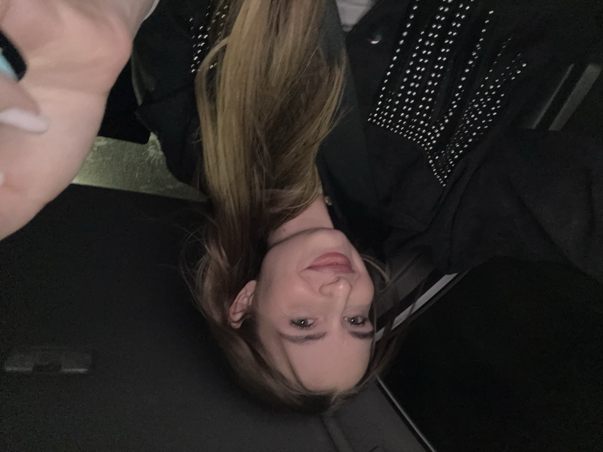 A woman is facing the camera with her head upside down in an indoor environment. She has long, brown hair partially covering the left side of her face and is wearing a black jacket adorned with studs. The surrounding environment is dark. The location is uncertain; Budva, Montenegro cannot be confirmed from the provided image.