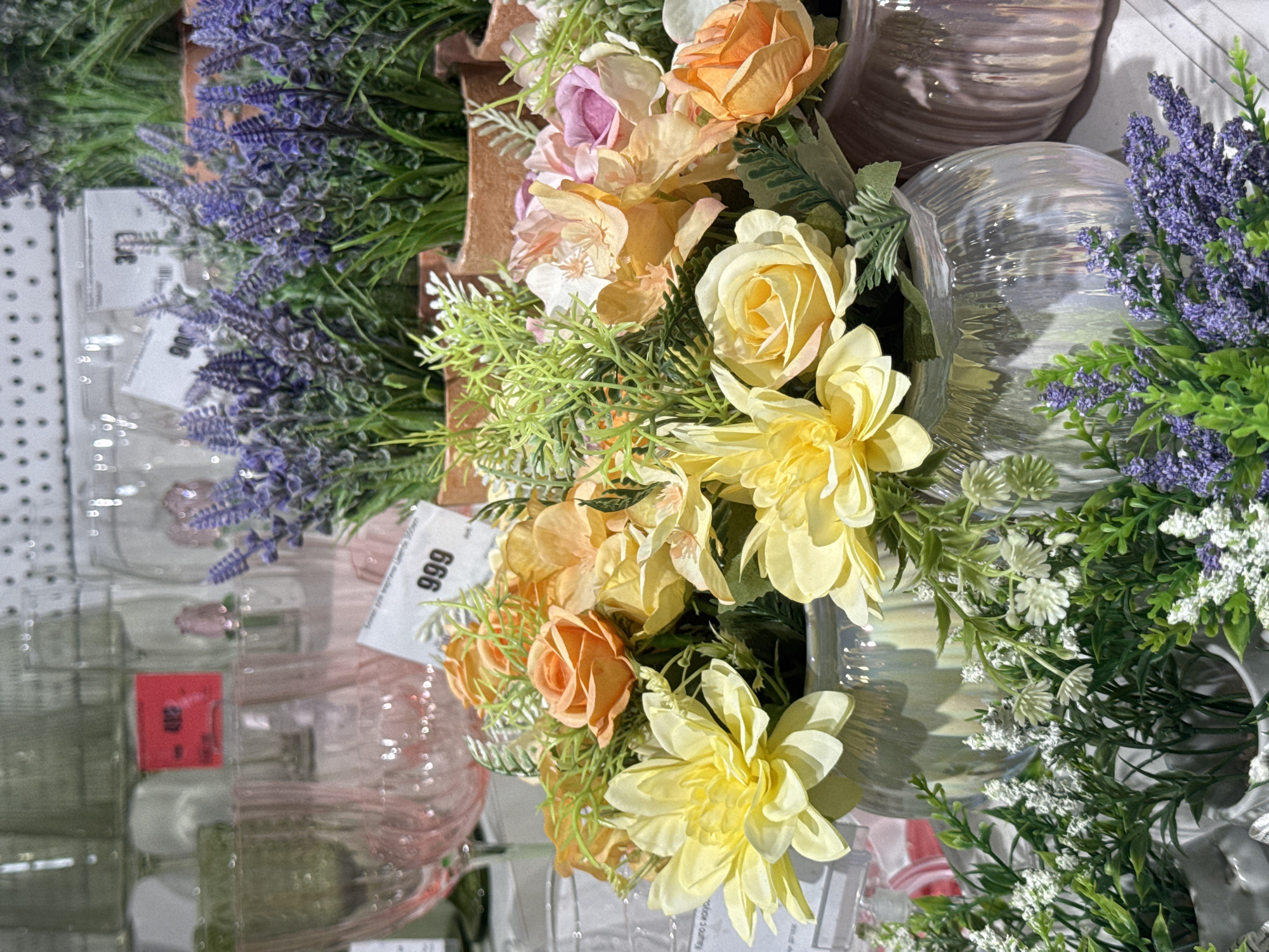 The image depicts an indoor display of artificial floral arrangements and glassware. In the foreground, several vases hold bouquets of artificial flowers in shades of yellow, orange, and pink, interspersed with green foliage. Some of the flowers appear to be roses and dahlias. Behind the flowers, several glass vases are visible, some with a pearlescent sheen. In the background, more artificial lavender plants in terracotta pots are displayed. There are also stacks of glassware, possibly wine glasses or goblets, some of which are tinted pink. Price tags are visible on some of the items, with one prominently showing the number "999". The overall setting suggests a retail environment, likely a home decor or flower shop. There is no indication of the specific location within the city of Ufa, Russia, or any people present.