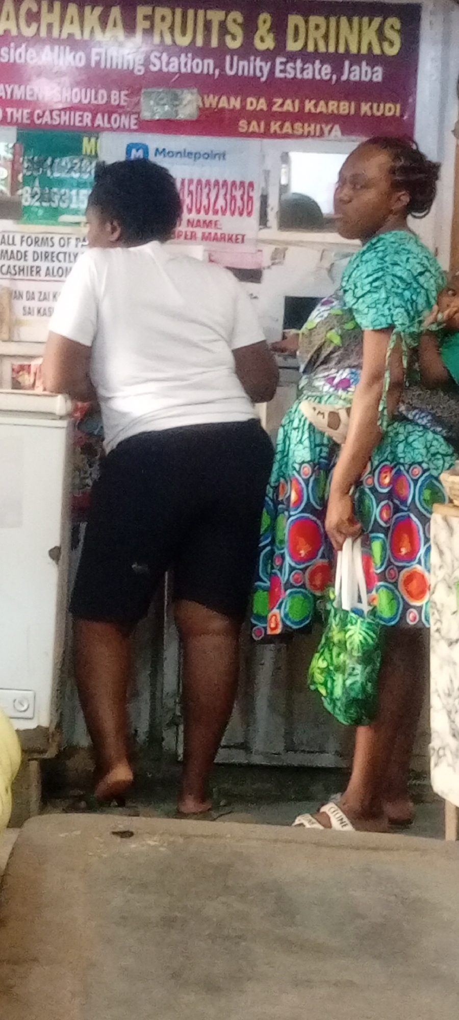 A photograph taken at a retail establishment called “Achaka Fruits & Drinks” in Kano, Nigeria. This shop appears to be located in the Unity Estate district of Jaba, next to an Aliko Filling Station. A sign above indicates that payments should be made directly to the cashier. A Moniepoint advertisement displays the phone number 0450323636.
Two Black women are present; one wears a white T-shirt and black shorts, standing with her back to the camera. The second woman is dressed in a colorful printed dress with a green bag. She is also wearing Celine sandals.
