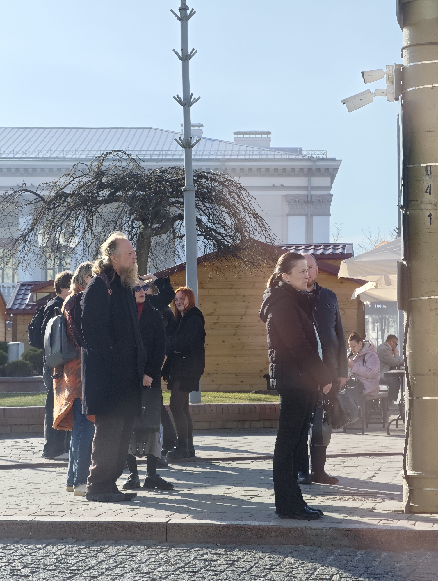The image depicts a group of people standing on a paved sidewalk. A man with a long beard is in the foreground, facing left. Beside him, several people are also looking in the same direction, including a woman with red hair. To the right of the frame, a woman in a black jacket stands with her hands clasped, looking towards something out of frame on the left. A man is beside her.
In the background, there are wooden structures, possibly stalls or small shops. Further back, a large building with a grey roof is visible. People are seated at tables outside a cafe. A metal pole is near the right edge of the frame, and it has security cameras attached to it.
The scene appears to be in Hrodna, Belarus.