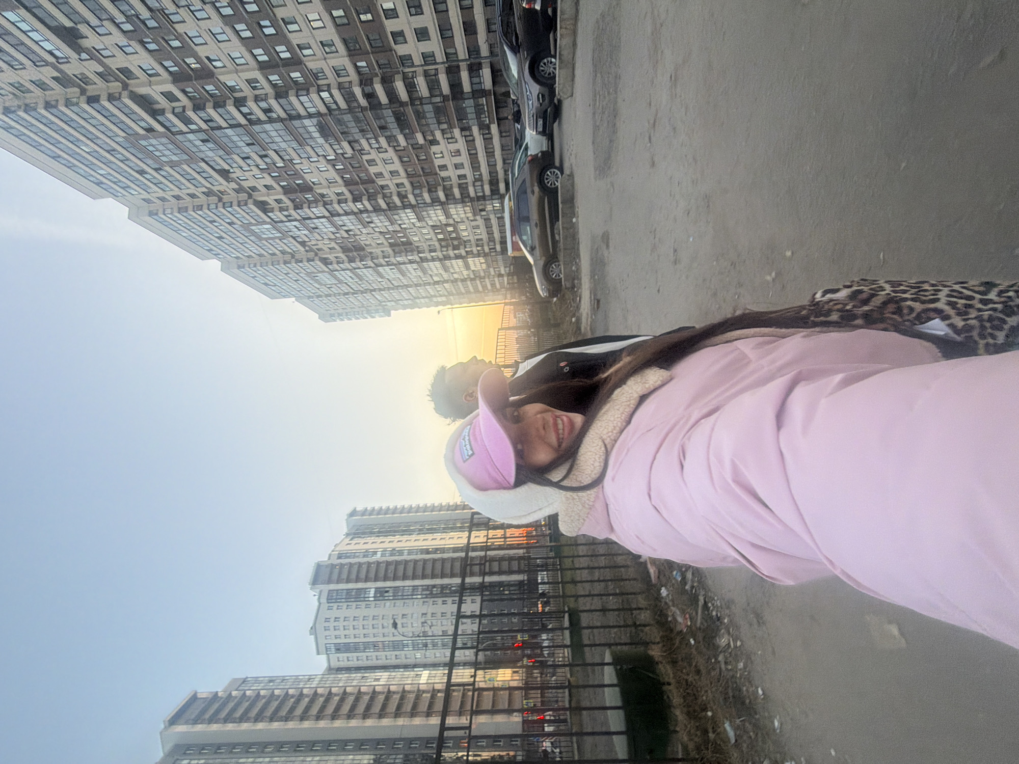 The image shows two people standing on a sidewalk next to a road in Saint Petersburg, Russia. The person closest to the camera is wearing a pink jacket and a pink hat. Behind them is another person with dark hair. Cars are parked along the road, and tall apartment buildings are in the background. The sky is bright with the sun low on the horizon.