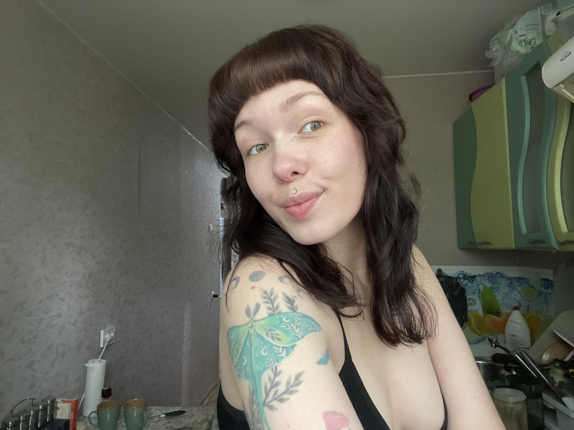 A woman is in a room in Chaikovsky, Russia. The woman has shoulder-length wavy brown hair with blunt bangs, fair skin, and green eyes. She is wearing a black top. She has a lip piercing on the right side of her lower lip. A tattoo of a moth, plants, and other designs is visible on her left shoulder. The room appears to be a kitchen, with patterned wallpaper and cabinets. There are cups and a kettle visible on a counter near a sink. A painting or printed image of lemons and oranges is on the wall near the sink.