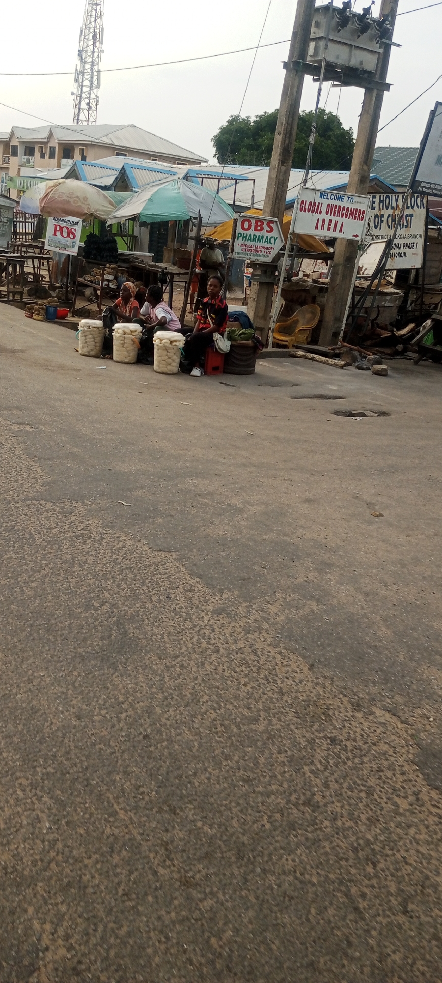 The image depicts a street scene in Lokoja, Nigeria. In the foreground, a paved road shows signs of wear with potholes and patches of rougher texture. On the right side of the road, a weathered utility pole with electrical equipment is prominent. Several signs are attached to or near the pole, including one that reads "WELCOME TO GLOBAL OVERCOMERS ARENA" and another for "OBS PHARMACY" listing services like "MEDICAL LABORATORY ULTRASOUND ECG".
Further back, a row of market stalls with colorful awnings is visible. Three women are seated by the roadside, in front of large, white, cylindrical containers, likely for sale or storage. One woman wears a headscarf, another has a pink top, and the third is in a patterned shirt. They appear to be vendors. Behind them, a larger building with a metal roof stands, and further in the background, a tall telecommunications tower can be seen. The overall impression is of a bustling roadside market in an urban environment.