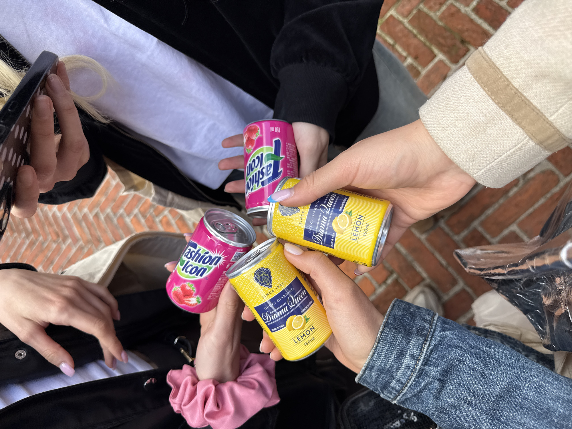 A high-angle, close-up shot captures the hands and arms of at least four individuals gathered outdoors in Milan, Italy. The focus is on three small beverage cans and a smartphone being held.
Two of the people are holding identical yellow cans, prominently displaying the brand "Gucci Giardino Drama Queen," featuring "LEMON" flavor and described as a "SPARKLING BEVERAGE." The cans also state "SINCE 1921," "MILANO ITALIA," and a volume of "150ml," with "GUCCI GIARDINO" repeated in a subtle background pattern. Another person holds a pink can labeled "Fashion Icon" with a strawberry graphic and "150 ml." A QR code is also visible on this pink can. A fourth hand is holding a black smartphone with a textured case.
The visible clothing includes a black long-sleeved top or jacket, a beige/cream jacket with tan trim, and a blue denim jacket. A white fabric, likely from a shirt or a bag's lining, is also seen, along with part of a white canvas bag. One wrist features a pink scrunchie. The hands show manicured nails, some with light pink polish and one with a blue French tip.
The background consists of red brick paving, indicating an outdoor setting. The natural lighting suggests the scene takes place during the day. The gathering appears to be a casual social event, with individuals sharing drinks.