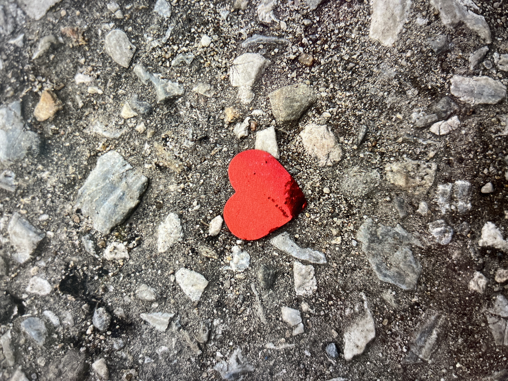 The image shows a textured gray surface composed of gravel, pebbles, and small rocks. In the center lies a small, red, heart-shaped object. The location is an unidentifiable outdoor area in an unknown city. No people are visible, and the scene depicts a single, stationary red heart on a rocky ground.