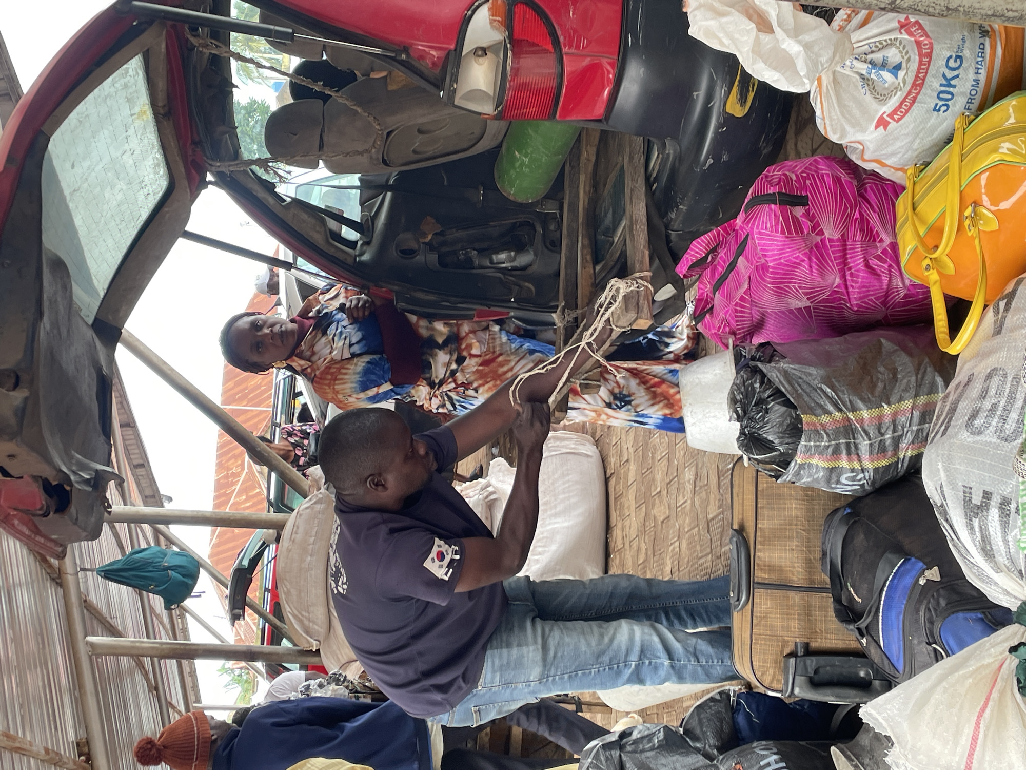 In Bukuru, Nigeria, a man in a dark blue t-shirt and jeans is seen tying a bundle of items with rope. He is bending over a wooden structure. Beside him, a woman in a colorful tie-dye shirt stands looking towards the camera. They are near the open rear of a red car, which is loaded with various items, including bags of produce, luggage, and a large green cylindrical object. Several other people are visible in the background, suggesting a busy marketplace or transport hub under a corrugated metal roof. The scene appears to be outdoors, with indications of tropical vegetation in the distance.