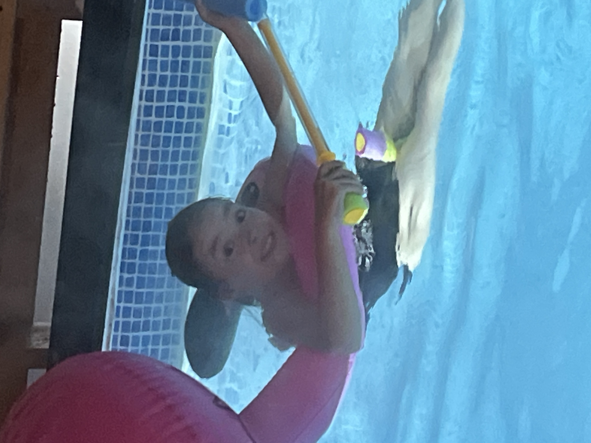 A young person wearing a pink swimsuit and riding a pink inflatable flamingo is partially submerged in a swimming pool with blue tiled walls. They are holding a water gun, with the barrel pointed upwards. Another individual is submerged further in the pool to the right of the young person, with their legs visible under the surface. The water in the pool is blue. The location and city are not discernible from the image.