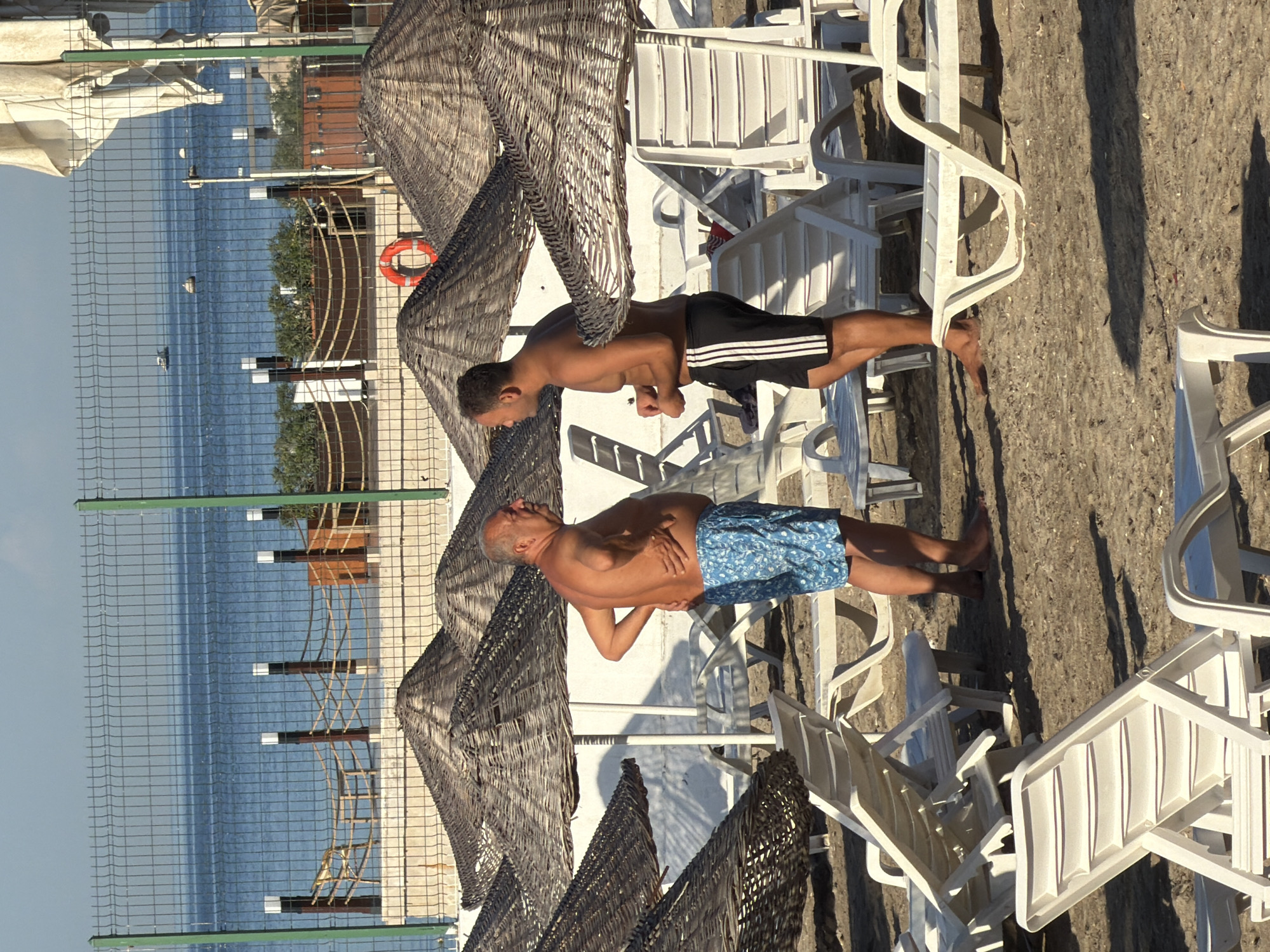 Two adult males are standing on a sandy beach in front of a wire fence. The male on the left is older, with gray hair and a fuller build, wearing blue patterned swim trunks. He is facing right, with his left hand on his lower back and his right hand touching his stomach. The male on the right is younger, with dark hair and a leaner build, wearing black athletic shorts with white stripes. He is facing left, with his hands clasped in front of him. Behind them are numerous thatched beach umbrellas and rows of white plastic lounge chairs. Beyond the fence, the blue sea is visible with a few small boats in the distance. The sky is clear and bright. The scene suggests a resort or public beach area, likely in Kadıköy, Turkey, given the context.