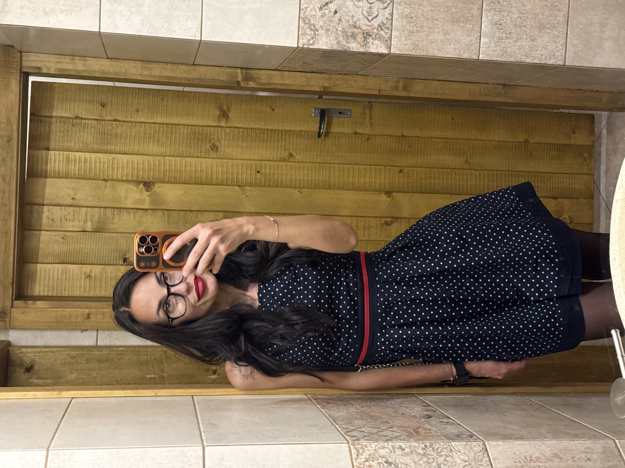 A woman with dark hair and glasses is taking a selfie in a room with tiled walls and a wooden door. She is wearing a dark blue dress with white polka dots and a red belt. The location appears to be indoors. I do not see information that provides the location in the city of Pleven, Bulgaria.