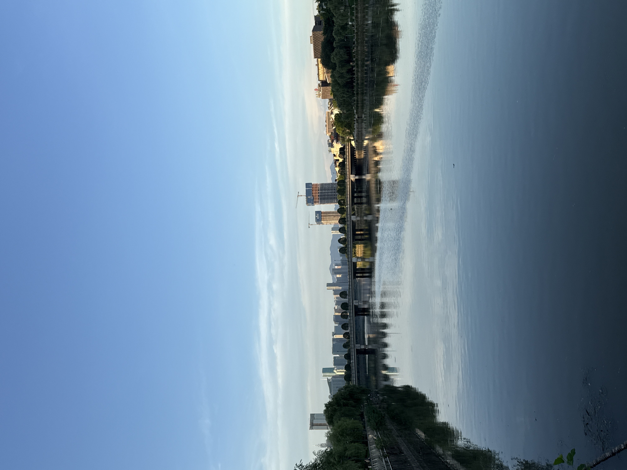 The image depicts a tranquil river flowing through the city of Yiwu, China. On the left bank, a tree-lined walkway with a stone embankment is visible, transitioning into lush greenery. Across the river, the Yiwu cityscape unfolds with a modern bridge spanning the water, supporting a road lined with evenly spaced trees and reflecting in the calm surface of the river. In the background, a cluster of high-rise buildings, some under construction with visible cranes, punctuates the skyline, interspersed with lower-rise structures and more trees. The sky is a clear, pale blue with faint wisps of clouds, suggesting it is either early morning or late afternoon, casting a soft, diffused light on the scene. The river itself is smooth, with clear reflections of the bridge, buildings, and trees, creating a symmetrical and serene panorama.