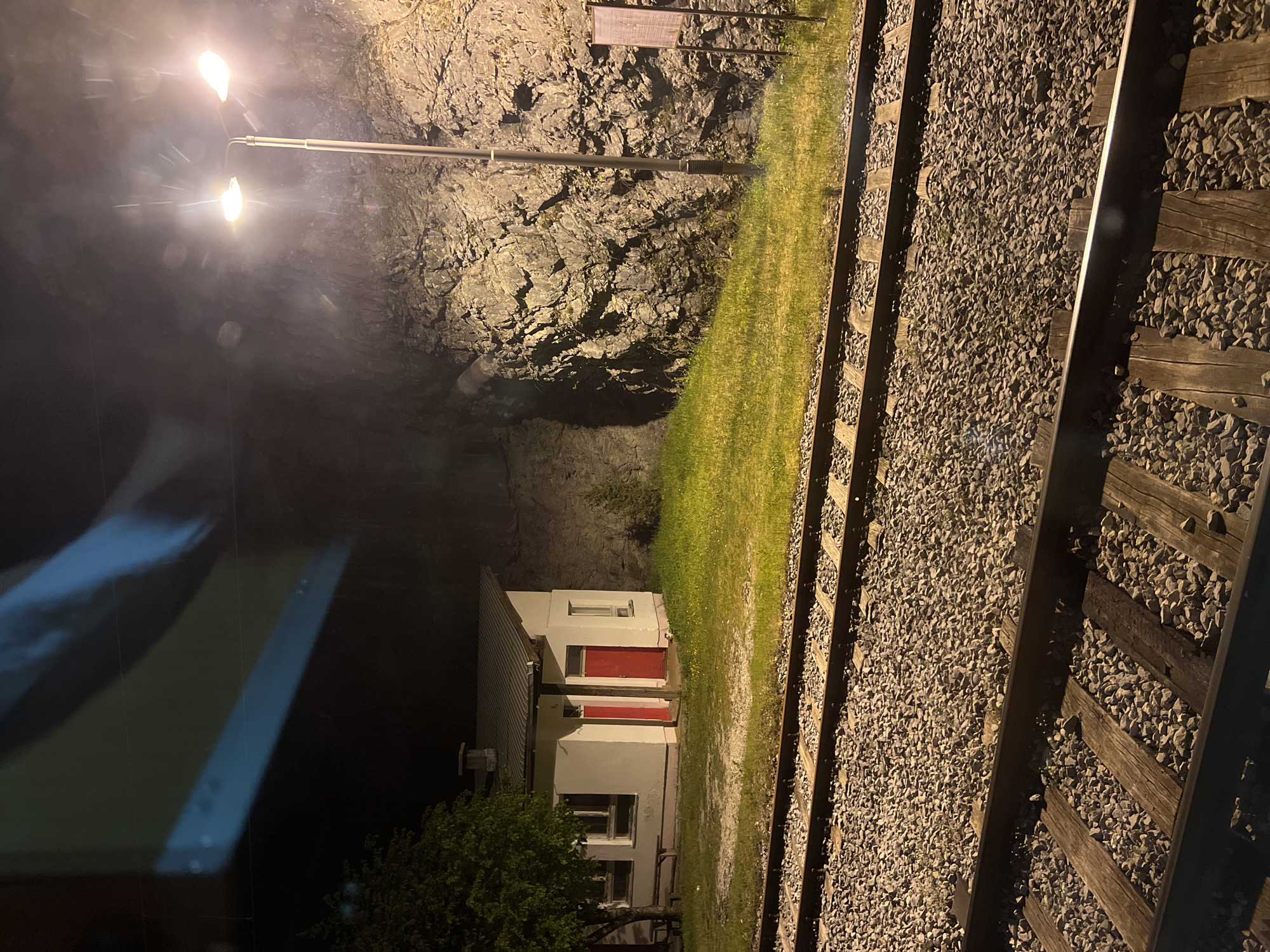 The image depicts a night scene alongside a railway track, likely in Lopate, Montenegro. The track is bordered by crushed gravel and wooden sleepers. Adjacent to the track is a strip of green grass. Beyond the grass, a small white building with red doors is visible. A rocky cliff face rises behind the building. A streetlight illuminates the scene. The left side of the image suggests the view is from inside a vehicle, due to the presence of a blurry reflective surface.
