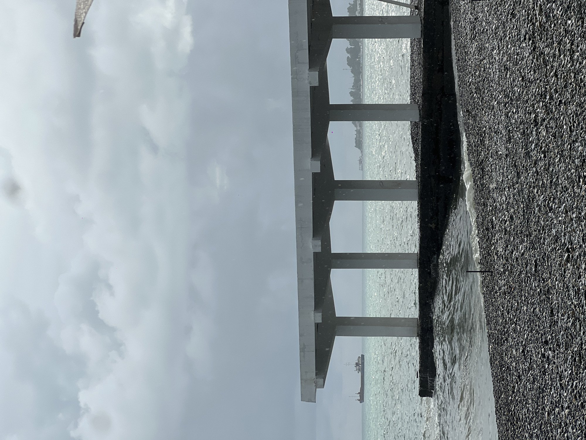 A concrete pier with multiple vertical supports extends over a pebble beach and into a choppy, grey sea under an overcast sky. A cargo ship is visible on the horizon in the distance. The weather appears to be raining, with visible streaks of precipitation in the air. The scene is devoid of people.